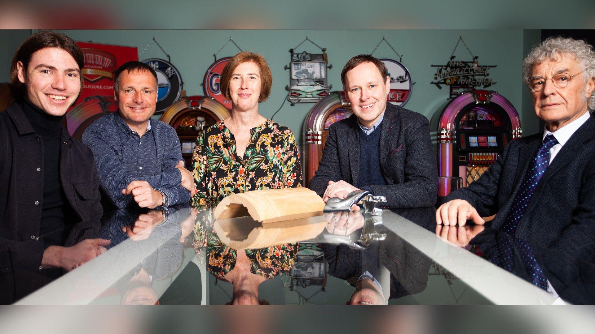 Four men and one woman sit around a board table with jukeboxes behind them.