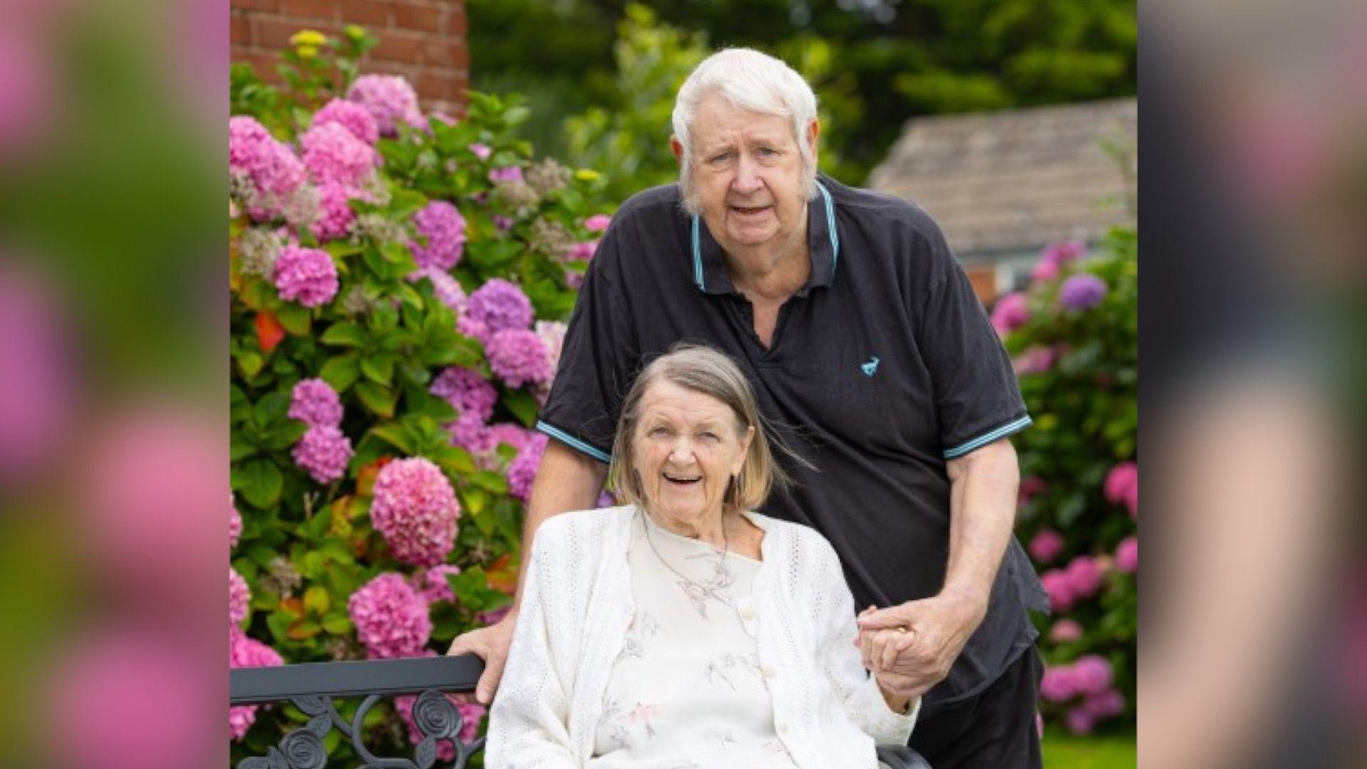 An elderly man and woman are in a garden surrounded by large pink flowers. The man, who has white hair and is wearing a black and blue-trimmed T-shirt, is standing and holding the hand of the woman who is sitting down. She has short fair hair and is wearing a white top and cardigan.
