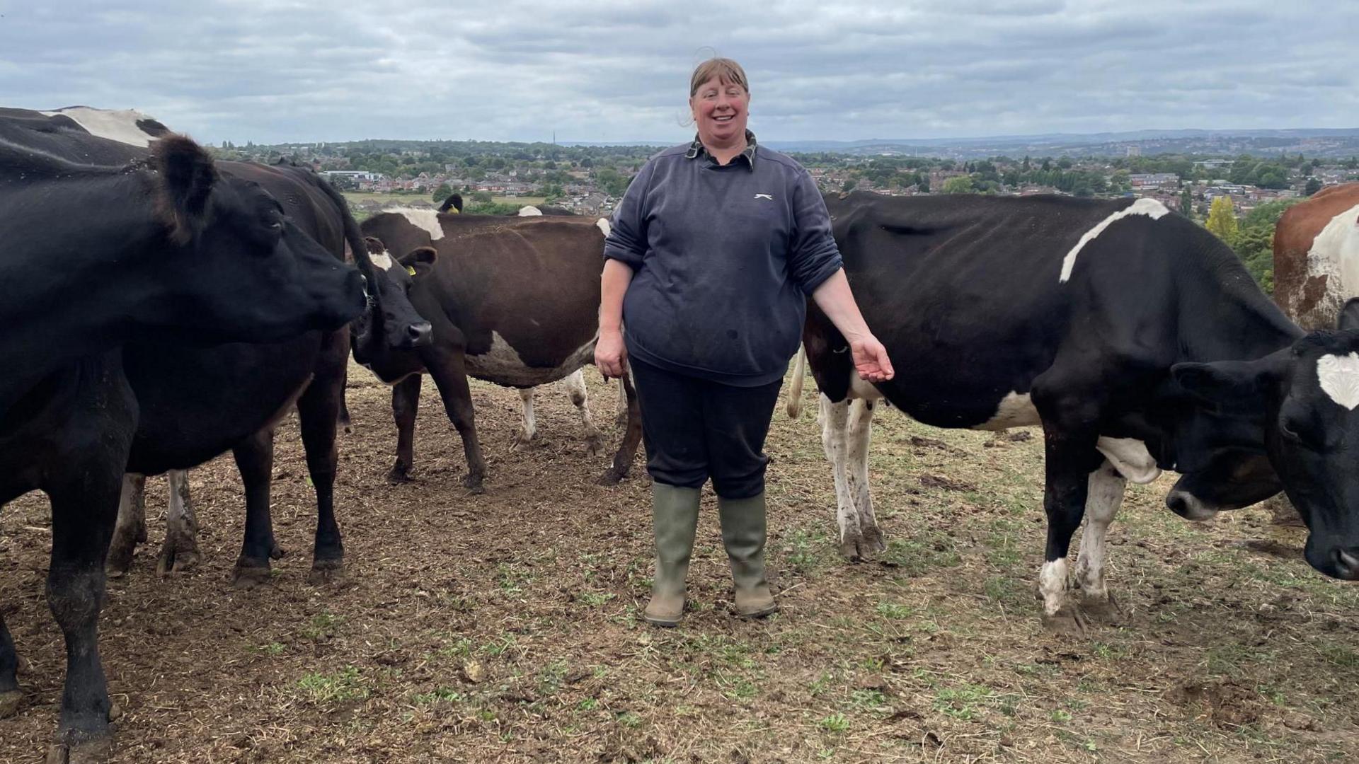 A woman in a comfortable navy jumper, black trousers and green wellies stands in a field surrounded by cows.