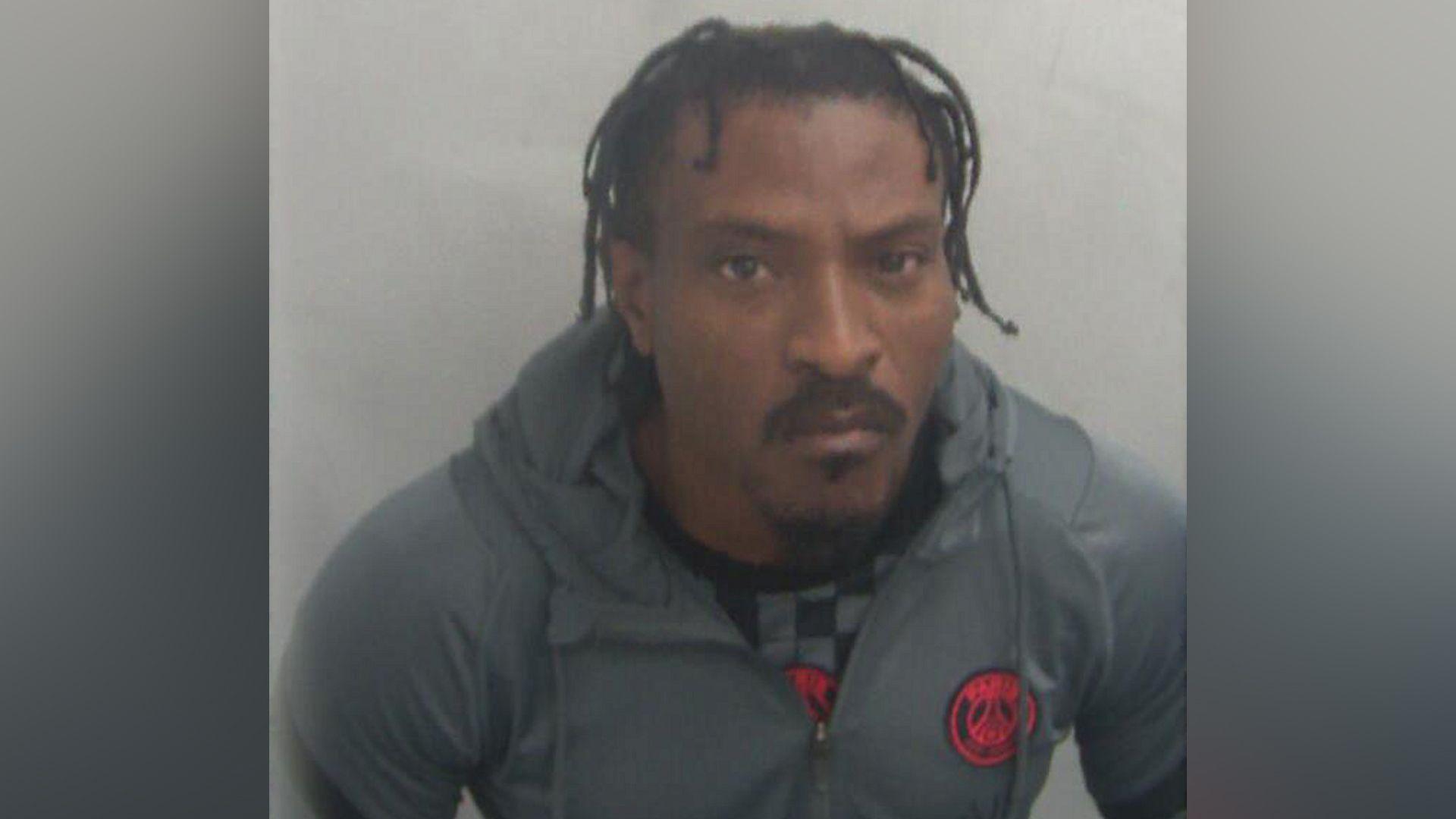 A police custody photo of Temitope Adeyinka. He has a stern look on his face. He has braided black hair, a moustache and a beard. He is wearing a grey hoodie with a grey and black chequered top underneath.