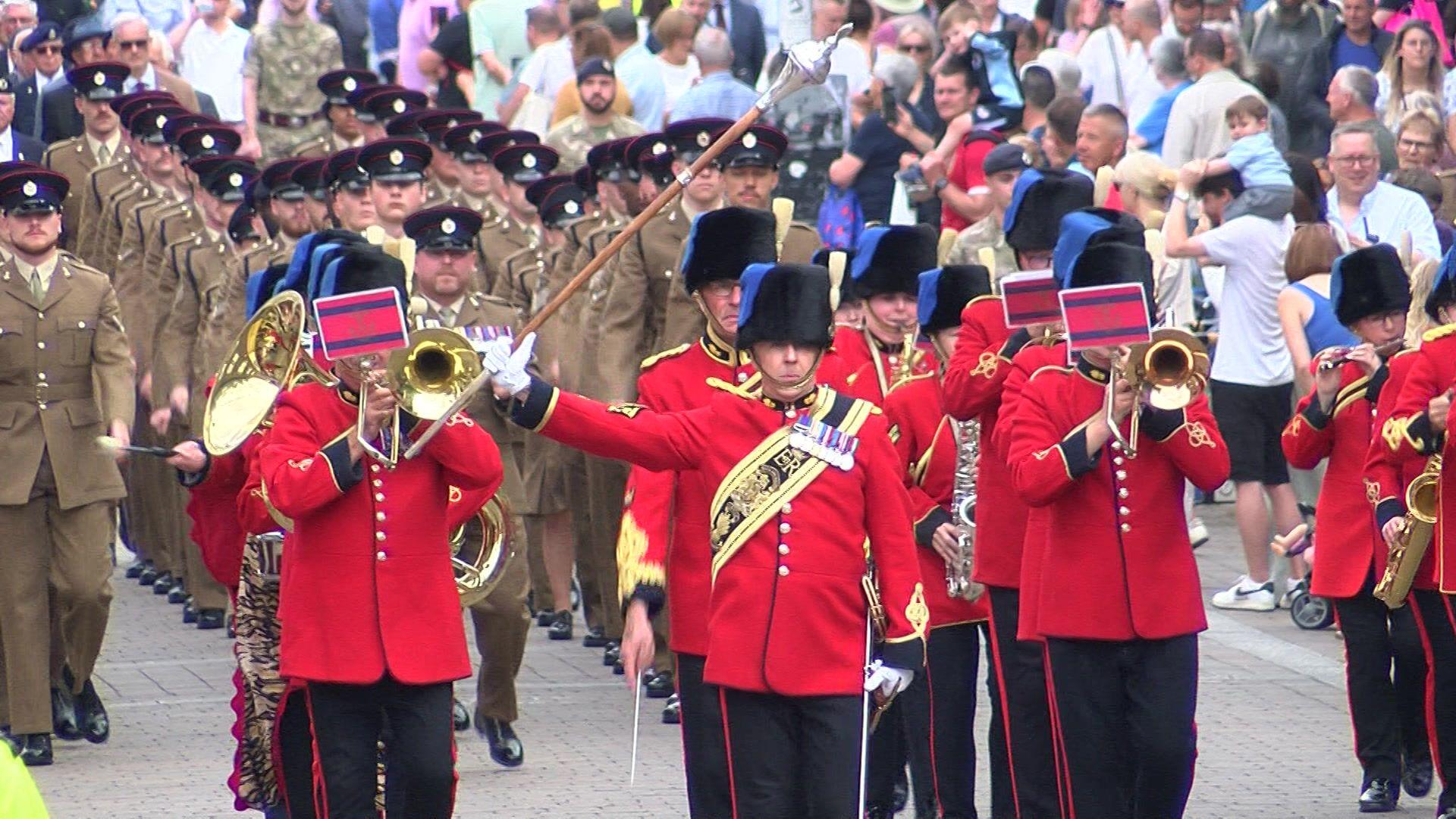 A large crowd surrounds columns of marching soldiers in brown uniforms in Newbury. At the front of the parade, a military band in red jackets and tall, black hats is led by a man holding up an ornate staff.