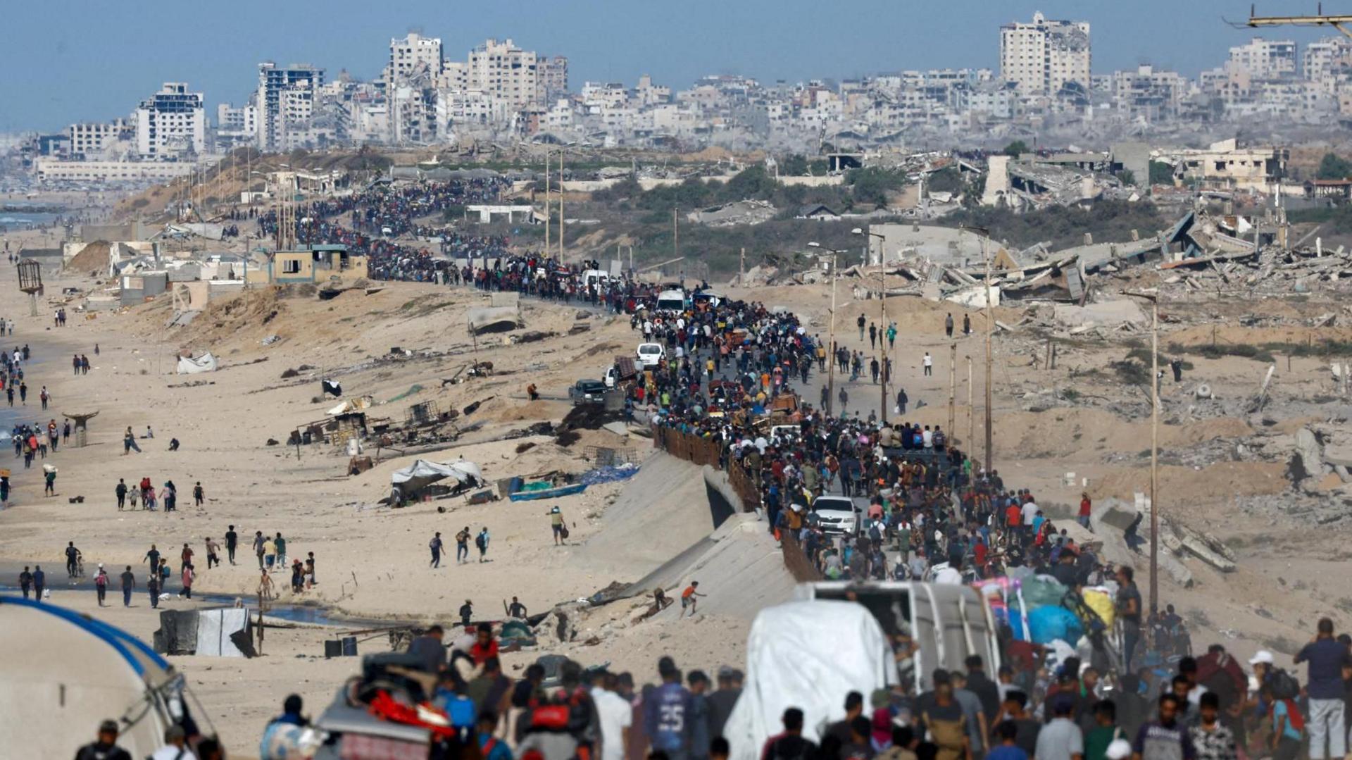 A long stream of Gazans walk along a road next to beach. Beside the road, and in the distance, there is debris and rubble from destroyed buildings.