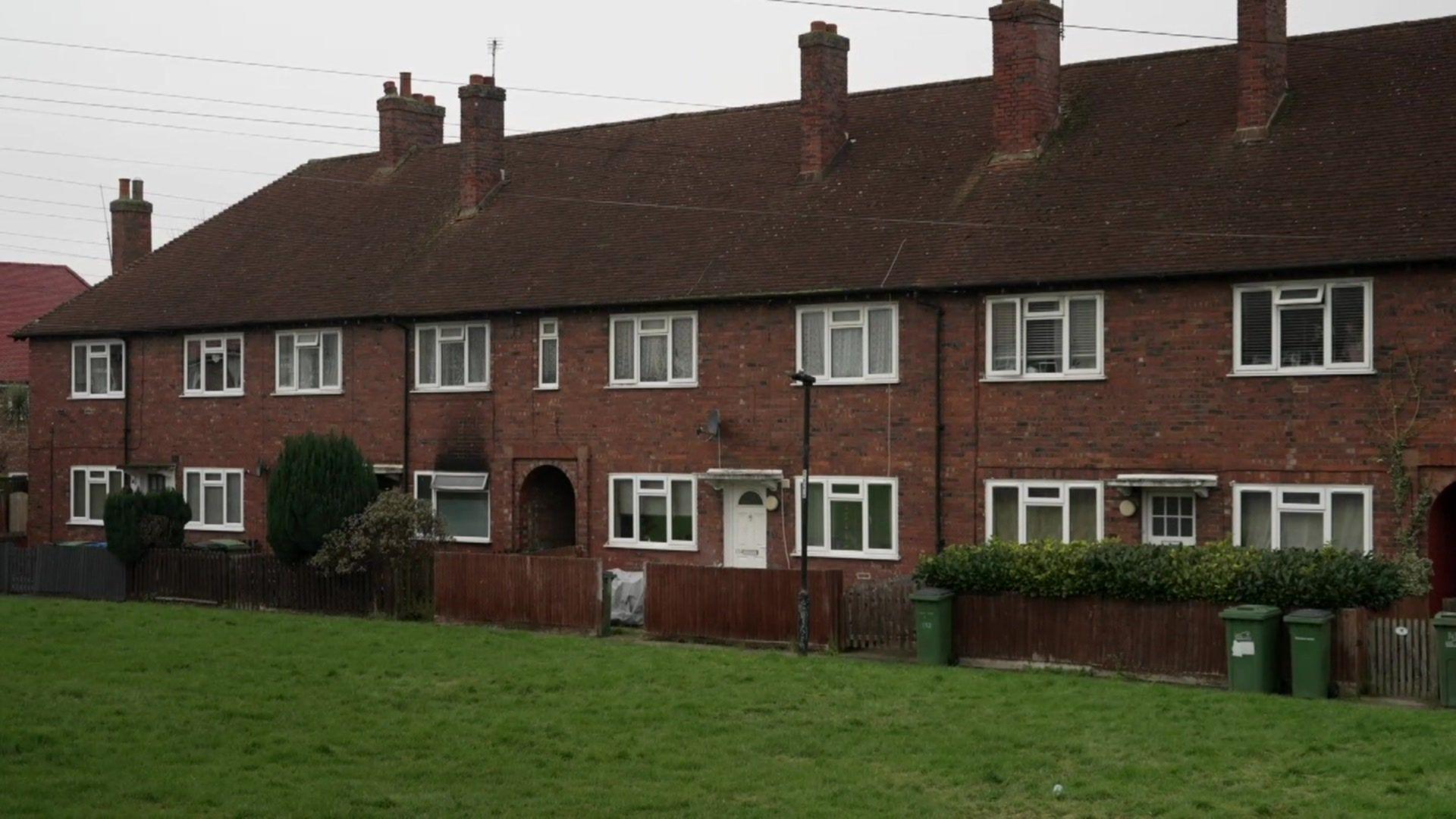 A row of red brick terraced houses with pitched roofs and multiple chimneys,. Each house has white-framed windows, small front gardens enclosed by wooden fences, and green wheelie bins. The foreground features a grassy area.