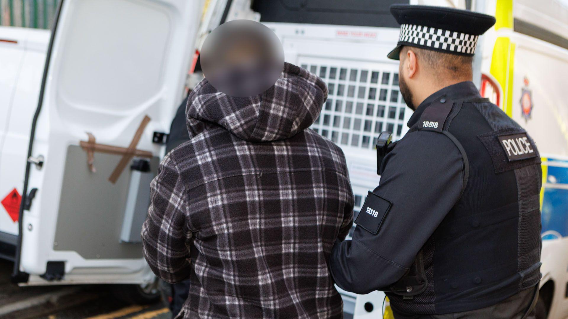 A police officer in uniform holds the arm of a man in a dark purple and white chequered hoodie as he guides him to the back of a police van. The suspect's face is blurred.