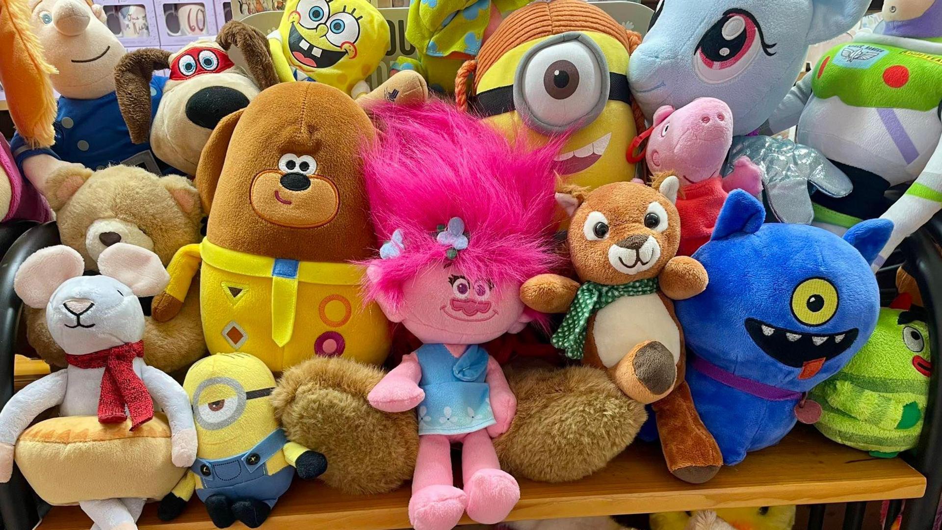 A set of soft toys lined up on a shelf in a card shop. They include a blue one-eyed monster, Gromit, some Minions, a pink troll, a mouse doing ballet and Fireman Sam.