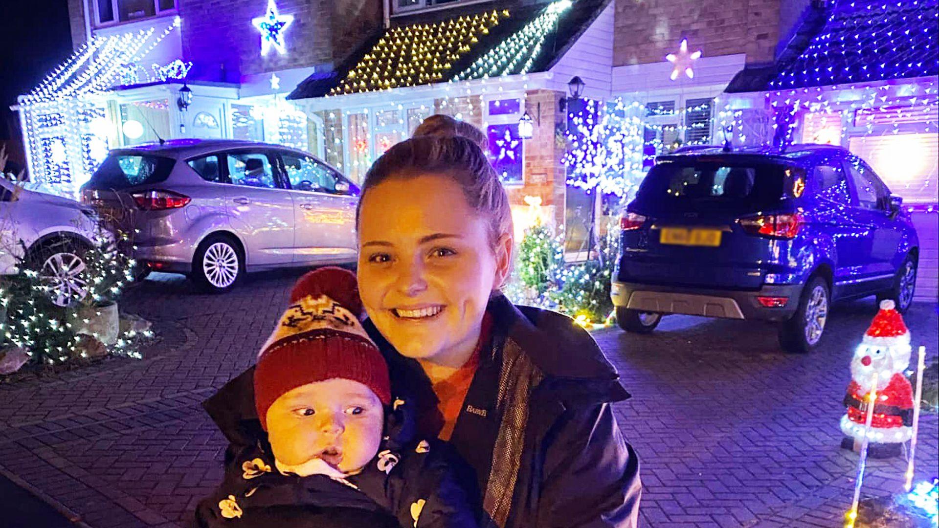 Stacey Newcombe holding her baby outside a house covered in lights. There are cars on the driveways. She has blonde hair and an anorak and the baby is wearing a red beanie hat.