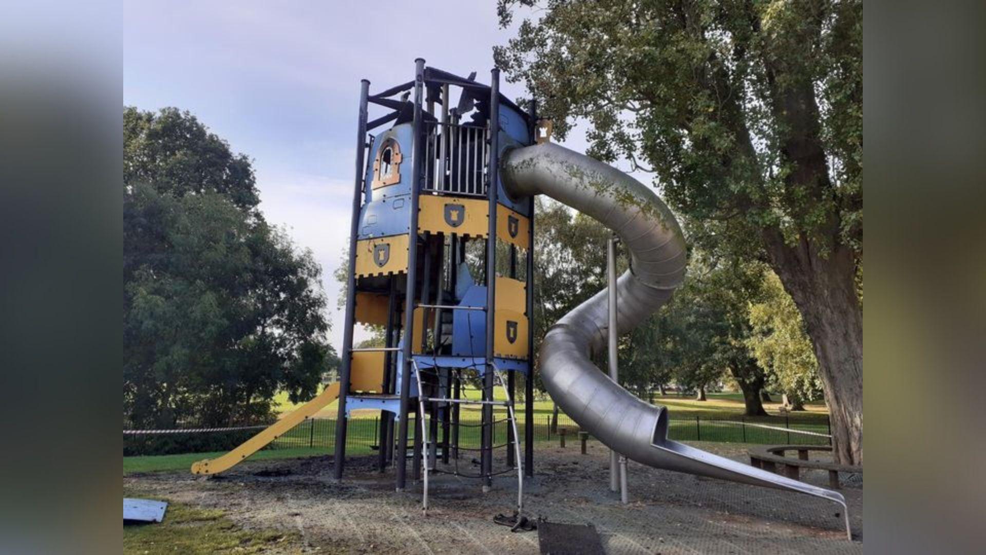 A large piece of playground equipment, painted yellow and blue, has been set alight and pieces are broken and burnt. 