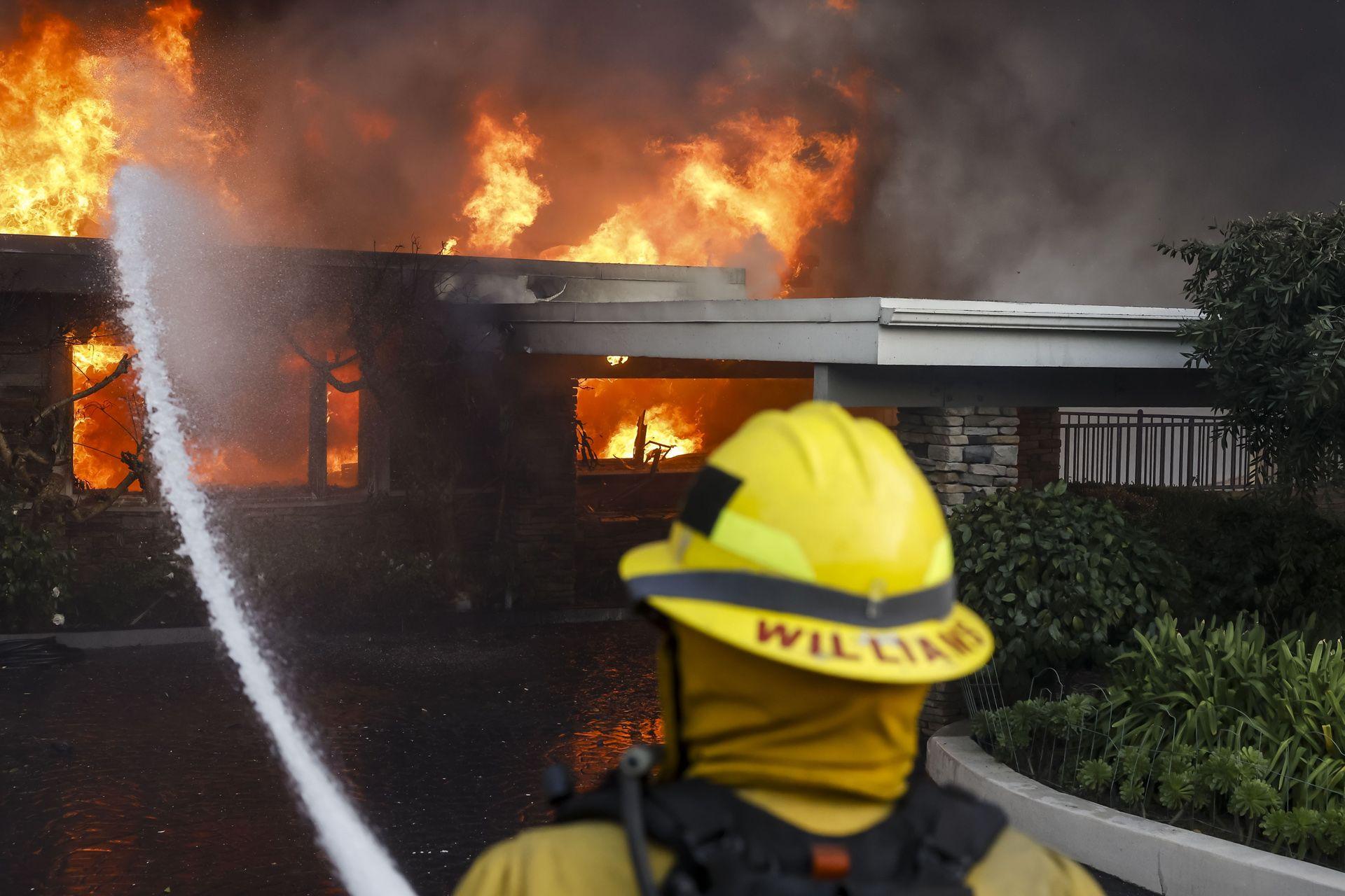 Rear view of a firefighter wearing a yellow helmet with the word 'Williams' on the back of it, as they fire a water cannon at a burning building in the Pacific Palisades.