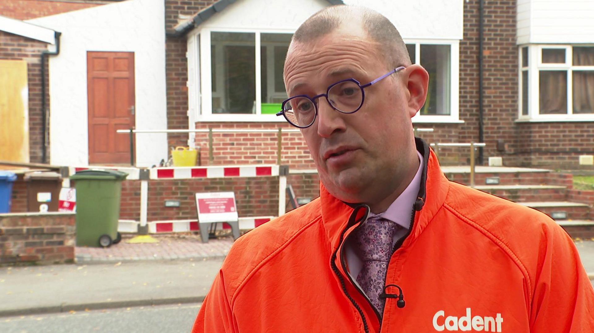 Dave Garner wearing orange Cadent jacket and black rimmed glasses in front of house undergoing building works