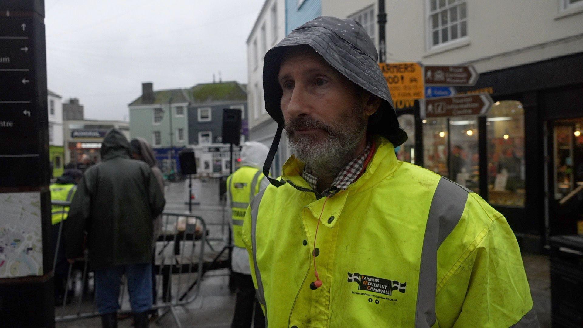 Peter Lawrence pictured in a high-vis jacket in front of the protest. Behind him people are standing, and there are signs on a signpost.