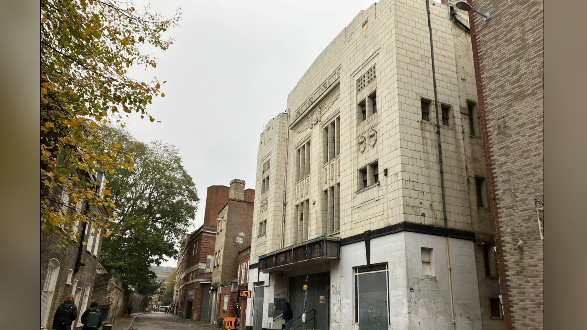 The old cinema seen on the right side of an old street with historical brick buildings on both sides. The lower part of the cinema is pale grey and dirty. There are the remains of a wooded hoarding above the door where the name would have been. The upper part is cream brick with art deco features, such as tall narrow windows. There are some trees on the opposite side further down.