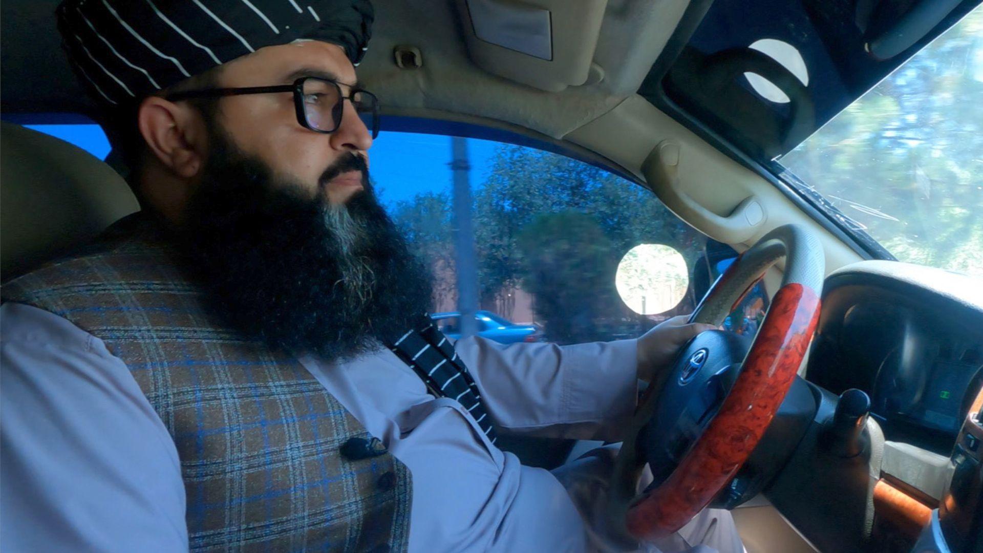Side shot of a man driving a car, photographed from the passenger seat. He has a full beard, glasses, black headdress with thin white stripes, a light blue top and checked waistcoat.