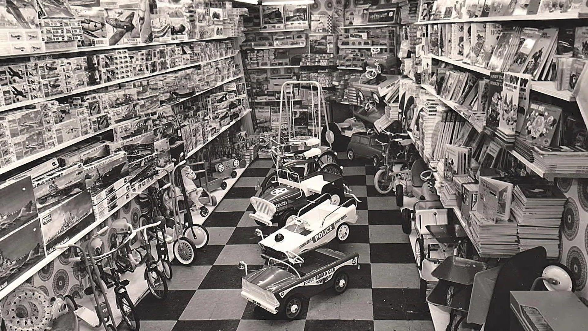 A black and white photo showing the inside of a toy shop with several bikes, puzzles, kits and toy cars