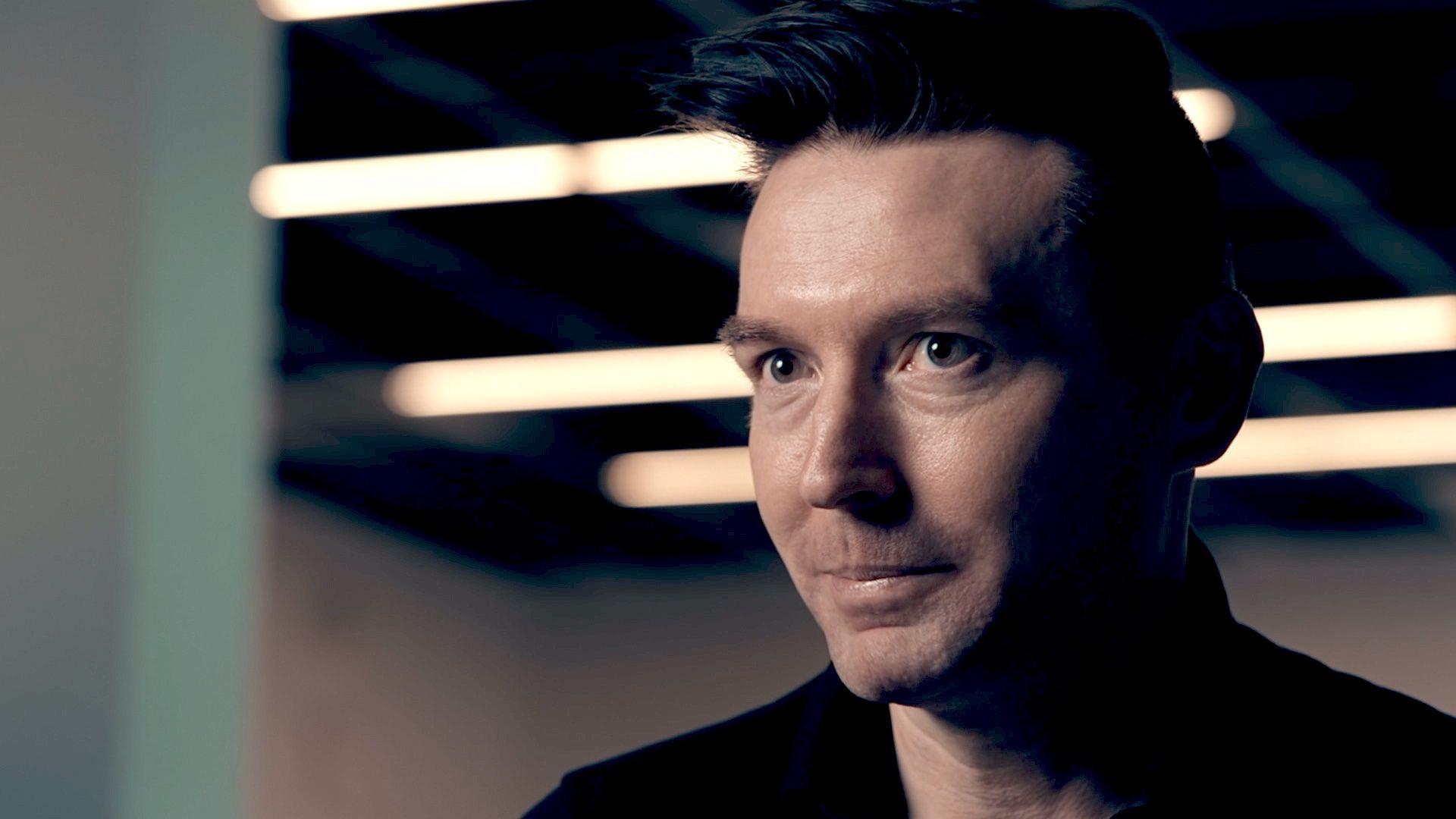 Matt Motyl, a man with brown hair styled in a quiff, pictured in close-up wearing a black shirt as he sits in an industrial-looking space with overhead strip lighting visible in the background blur.