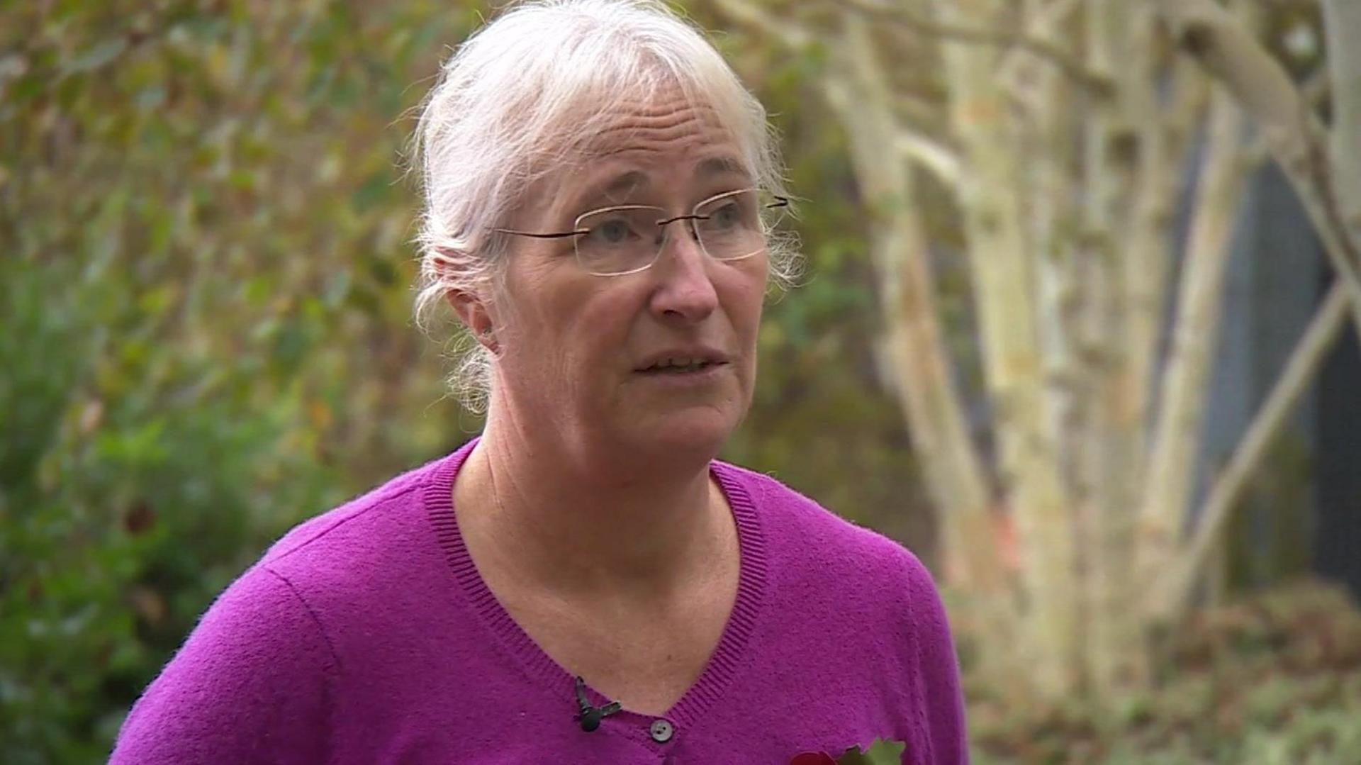 Debbie Moreton looks slightly off camera while being interviewed, there are trees and greenery slightly blurred in the background behind her. Debbie has white hair, tied back in a bun and a fringe framing her face. She has clear framed glasses and wears a magenta pink wool cardigan with black small buttons.