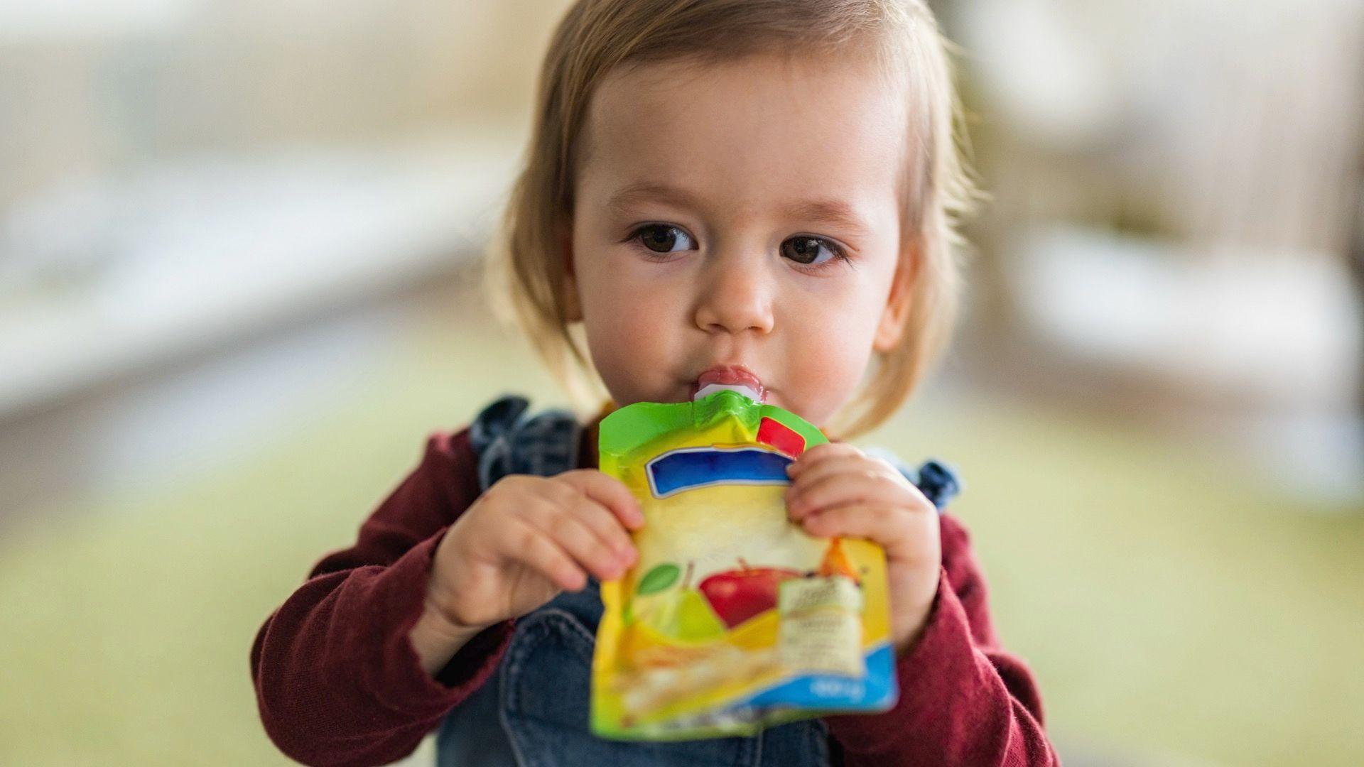 A toddler with blondey-brown hair sucking a pouch of baby food, which she is holding with both hands. She is wearing a burgundy top and dungarees.