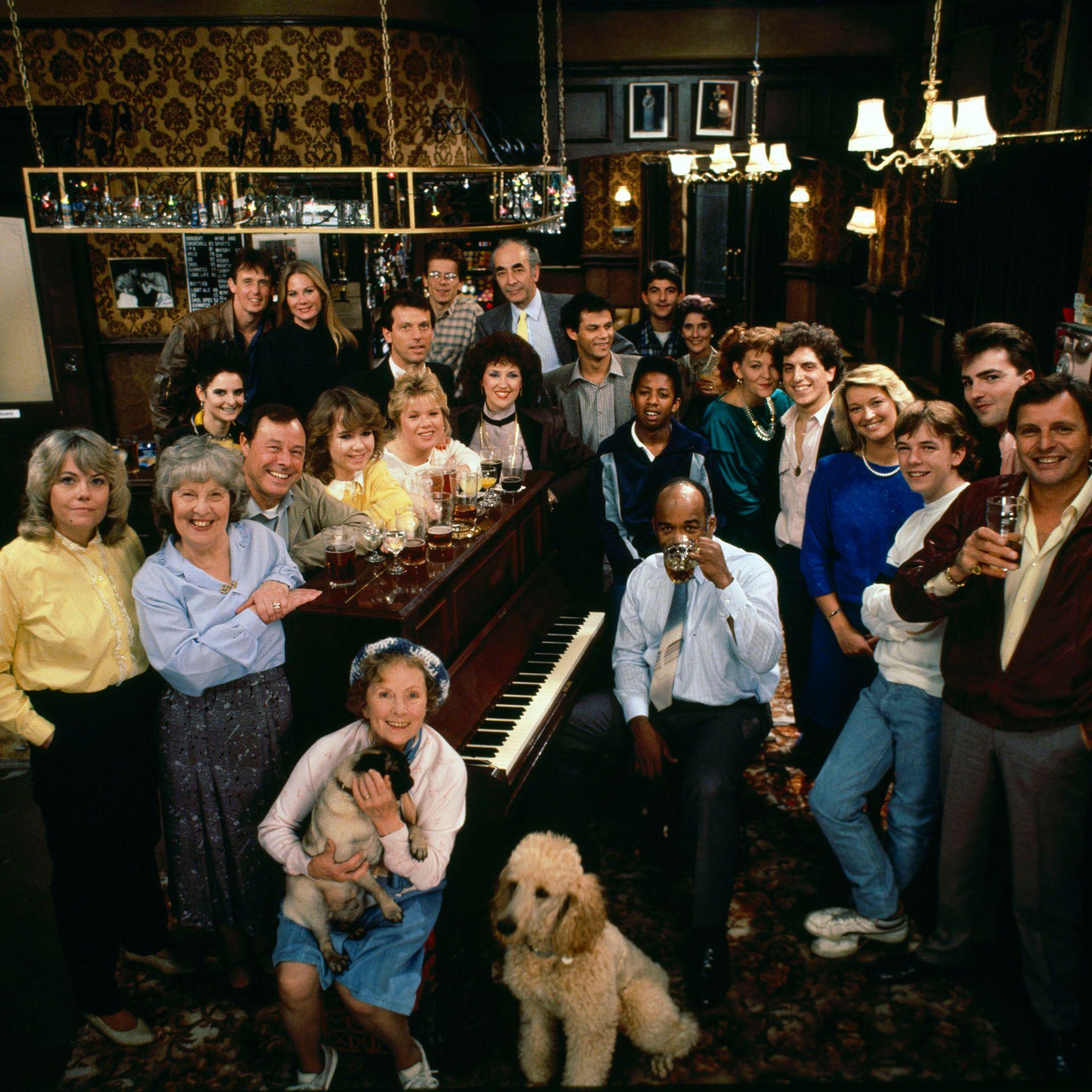 The cast of EastEnders in 1985, sat around the Queen Vic piano