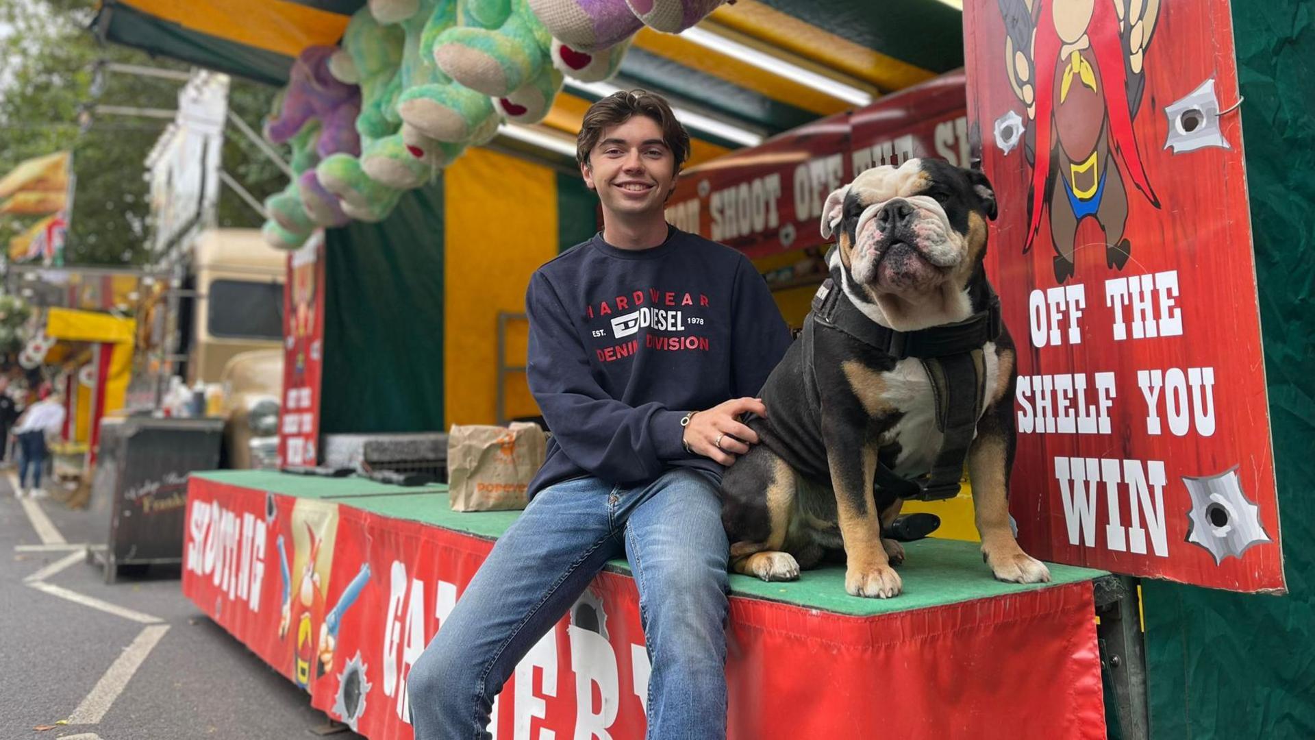 A young man sits with his dog in front of a Western-themed shooting range at the fair.