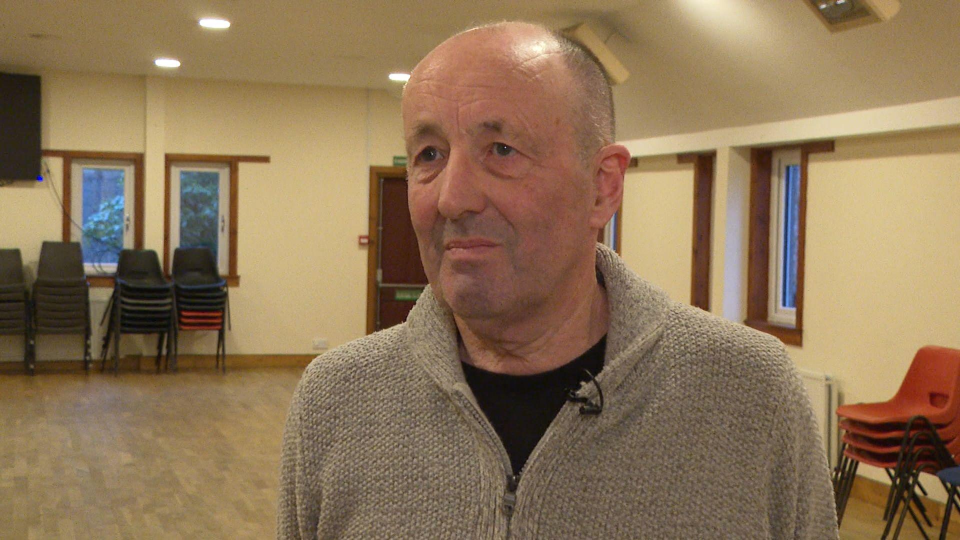 Ruairidh Peutan na sheasamh ann an talla