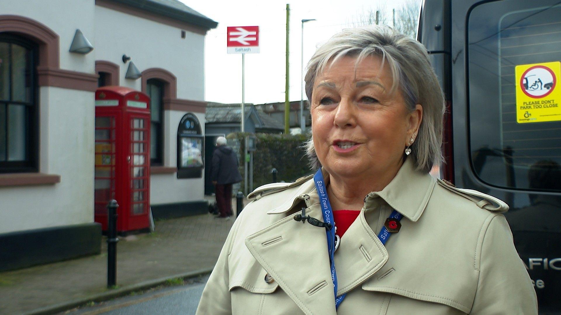 Julia is wearing a cream trench coat and is standing outside Saltash station with the railway sign and a red phone box behind her