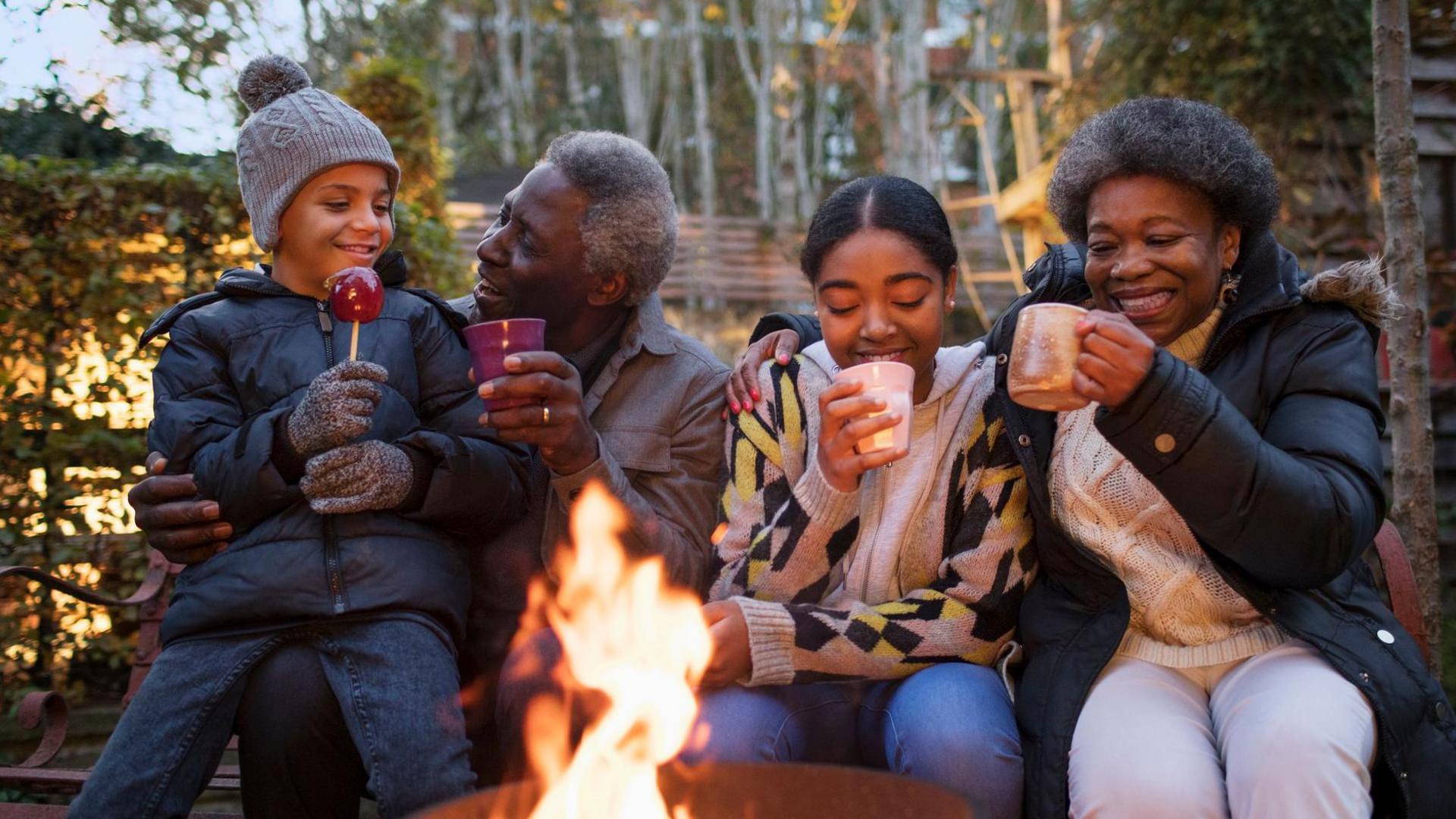 Grandparents and grandchildren drinking hot chocolate and toffee apples at campfire.