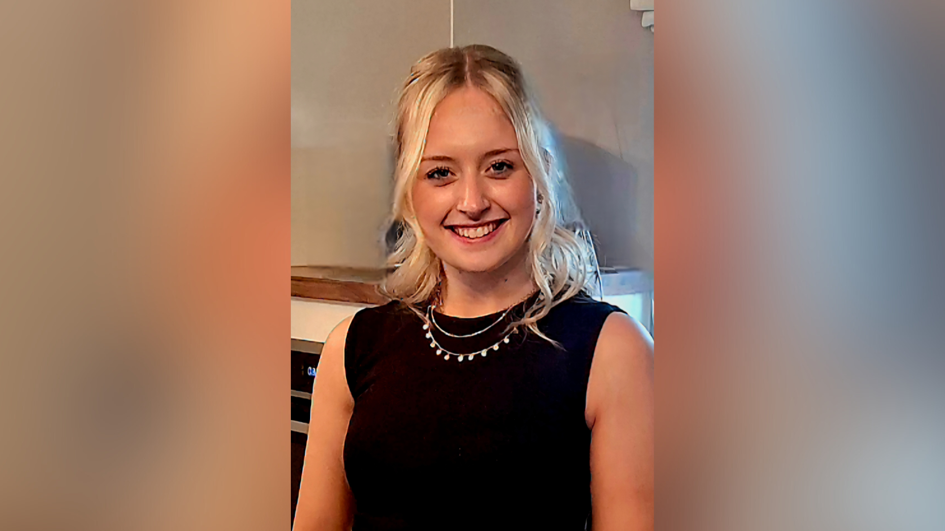 A young woman is wearing a sleeveless black top and a necklace, and has long blonde hair. She is smiling at the camera indoors.
