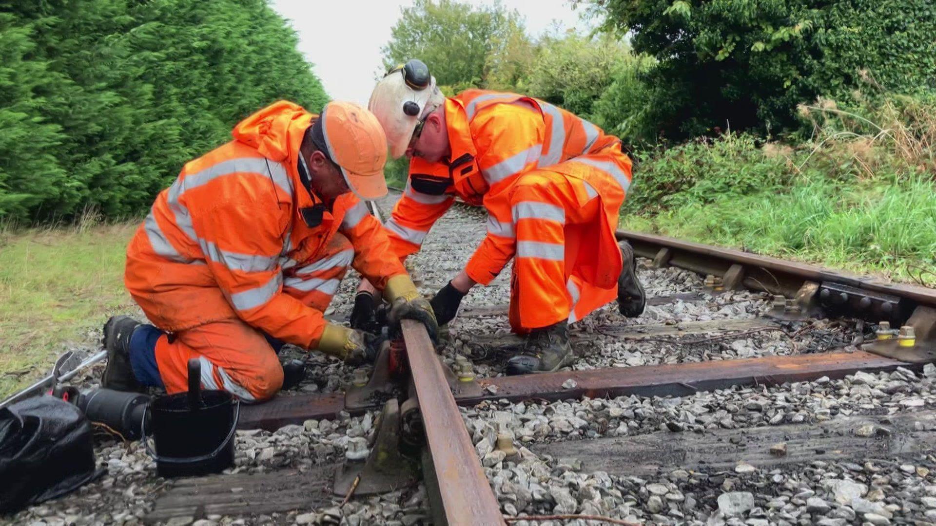 Two people wearing high-visibility orange protective clothing remove a bolt from a railway track, crouching on the line.