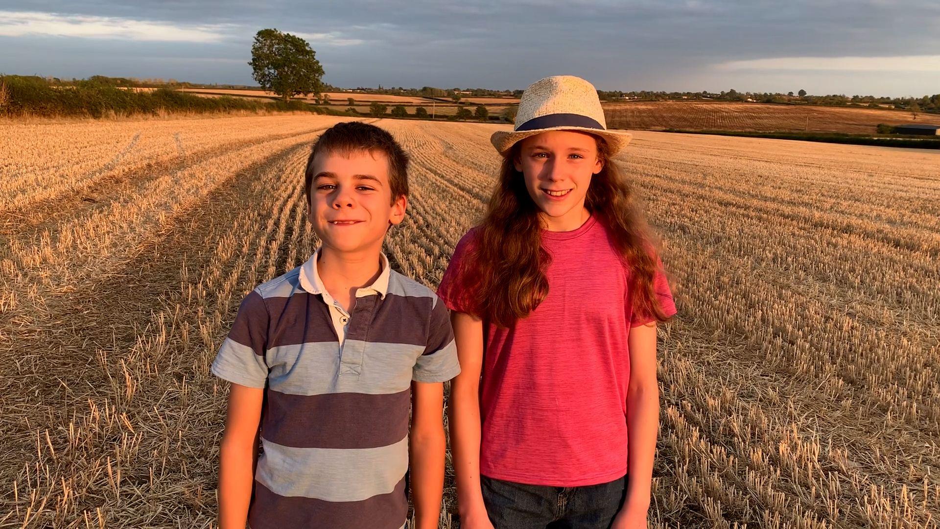 A boy with a blue stripy t -shirt and a girl with a pink t-shirt, long wavy hair and a hat standing in a field where teh crops have been cut. Both are smiling and looking straight at the camera.