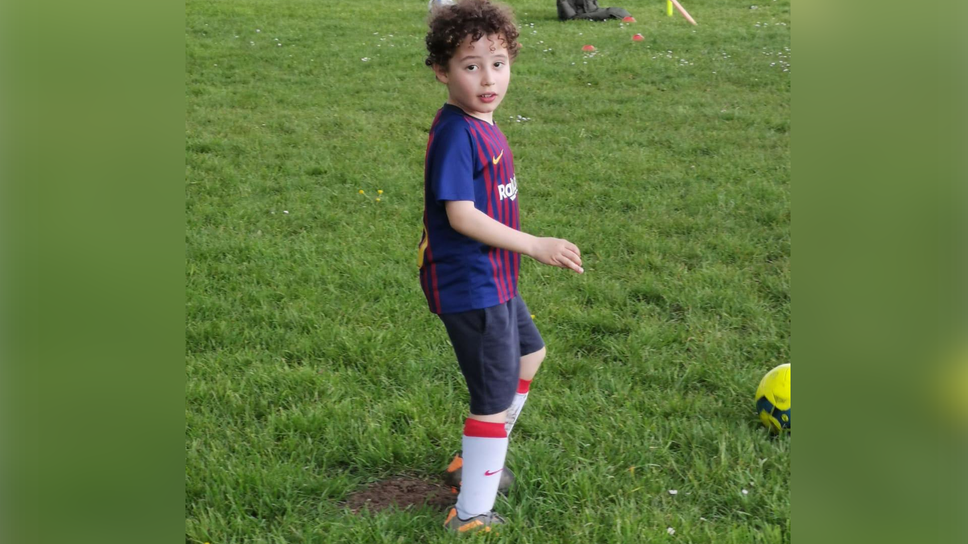 Young boy in a football strip. He is on a football field and there is a ball next to his foot. He is looking at the camera