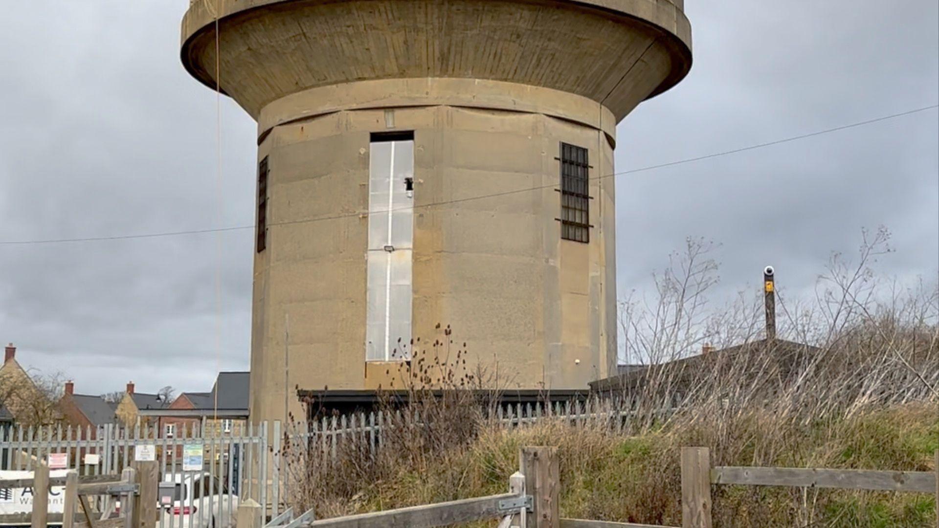 A long shot of the beige-stone water tower, which rises up behind a metal fence and some bushes.