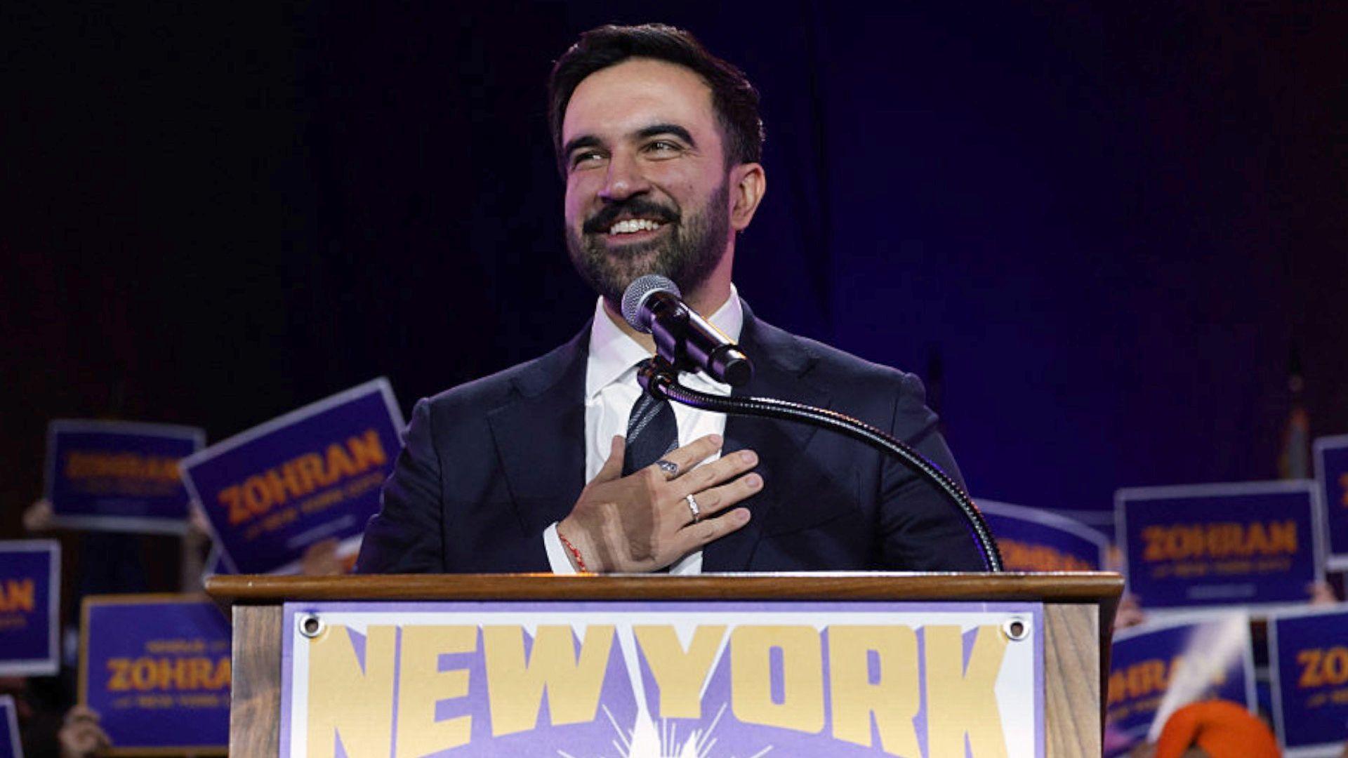 A man stands at a podium with his hand on his chest smiling - signs behind him say 'Zohran'