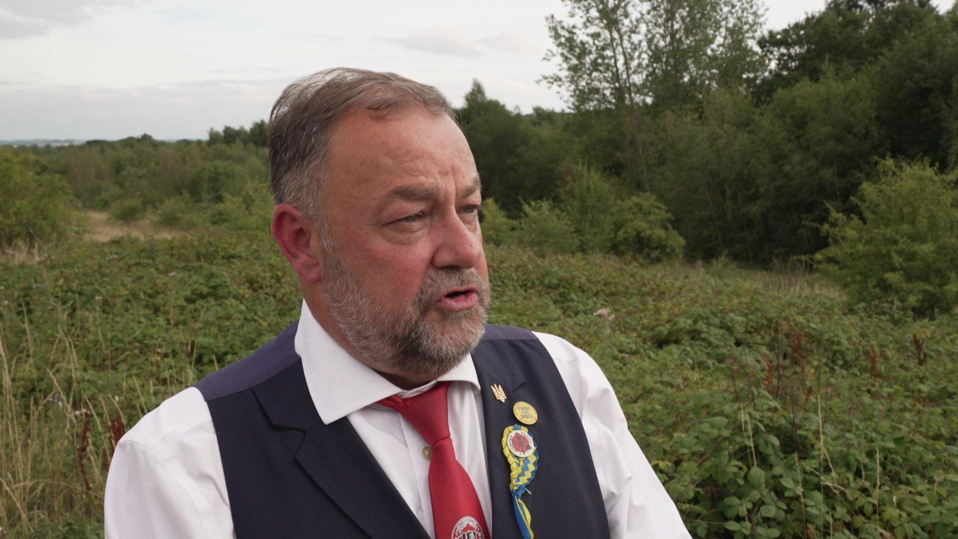 Chris Kitchen is stood outside and has short dark hair and a short beard. He is wearing a red tie, white shirt and black waistcoast, with some emblems on the lapel of the waistcoast