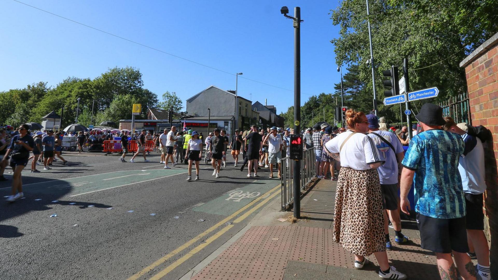 Oasis fans queue by the side of a wall that marks the boundary of Heaton Park, while others cross a road from the Metrolink stop to the park in the background
