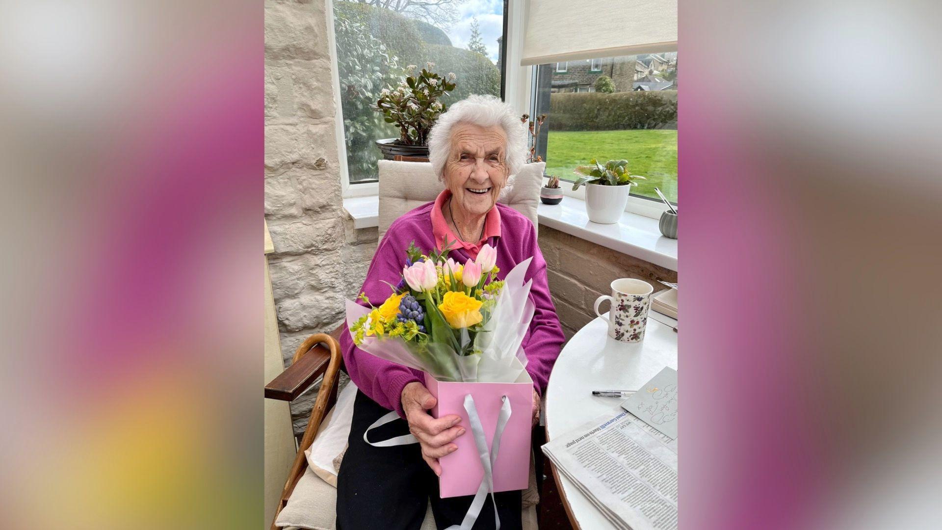A 98-year old woman is sitting in a conservatory holding a large bunch of flowers.  She is next to a table that has an empty coffee cup and a newspaper on it.  She is wearing a dark pink jumper and blue trousers.