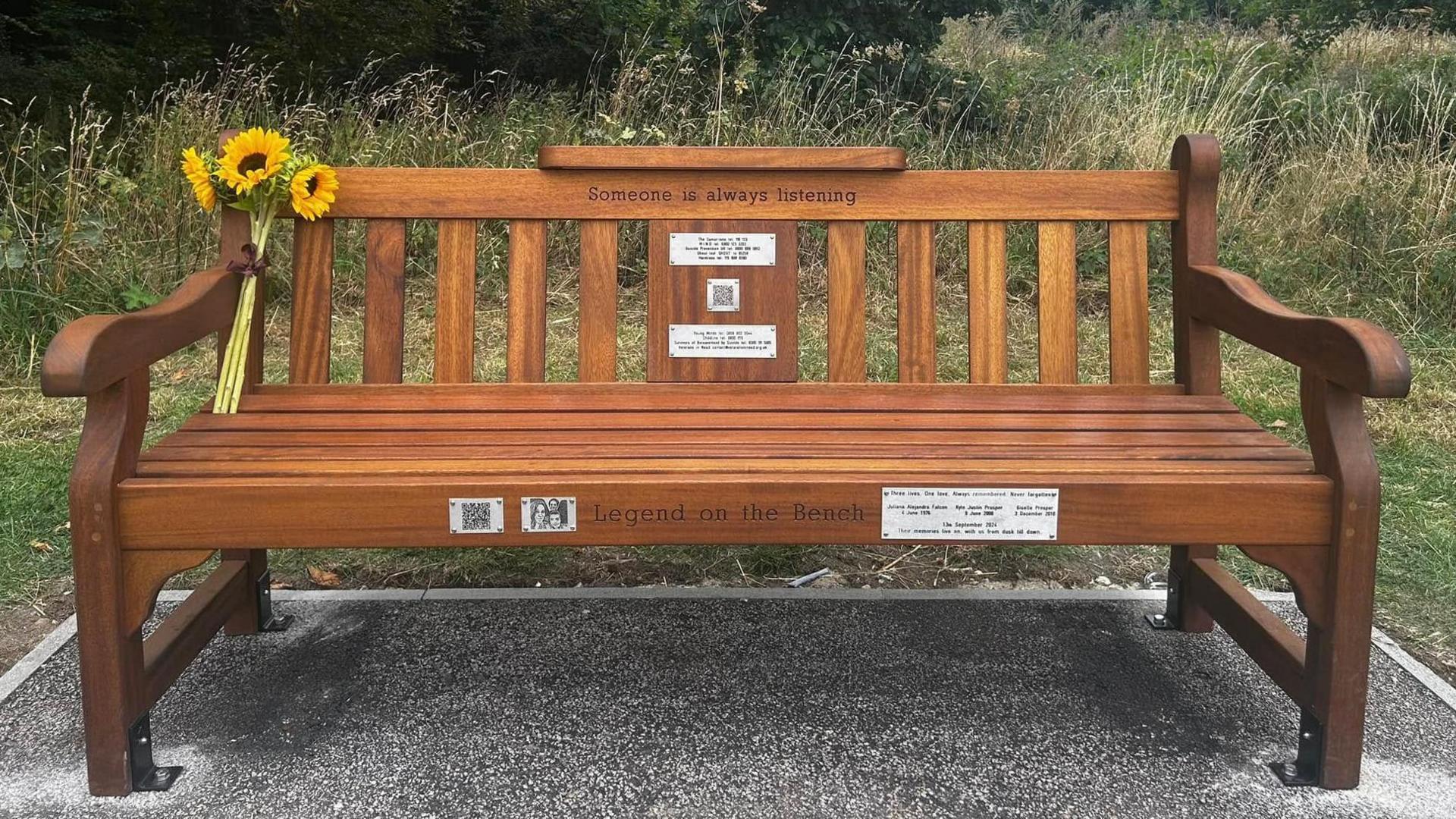 Wooden varnished bench with "someone is always listening" engraved on the top of the back panels. There are metal plaques with engraved writing on the back panel and front of the seat. A bouquet of sunflowers rests in the left back corner of the bend. It is standing on a stone plinth with grass and hedge behind.