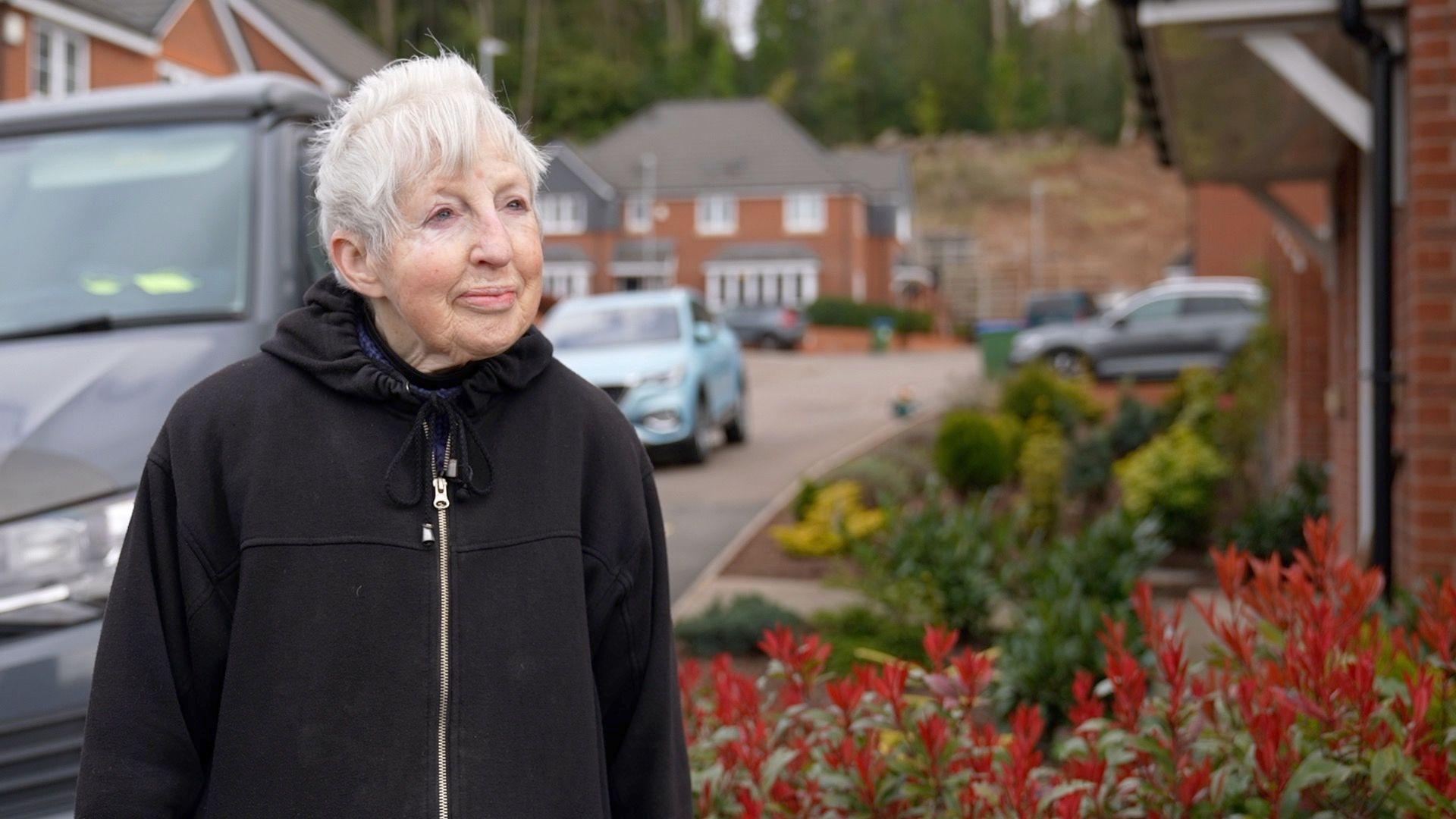 Irene Taylor is standing outside a home with a van and car on the left and plants outside the row of houses on the right