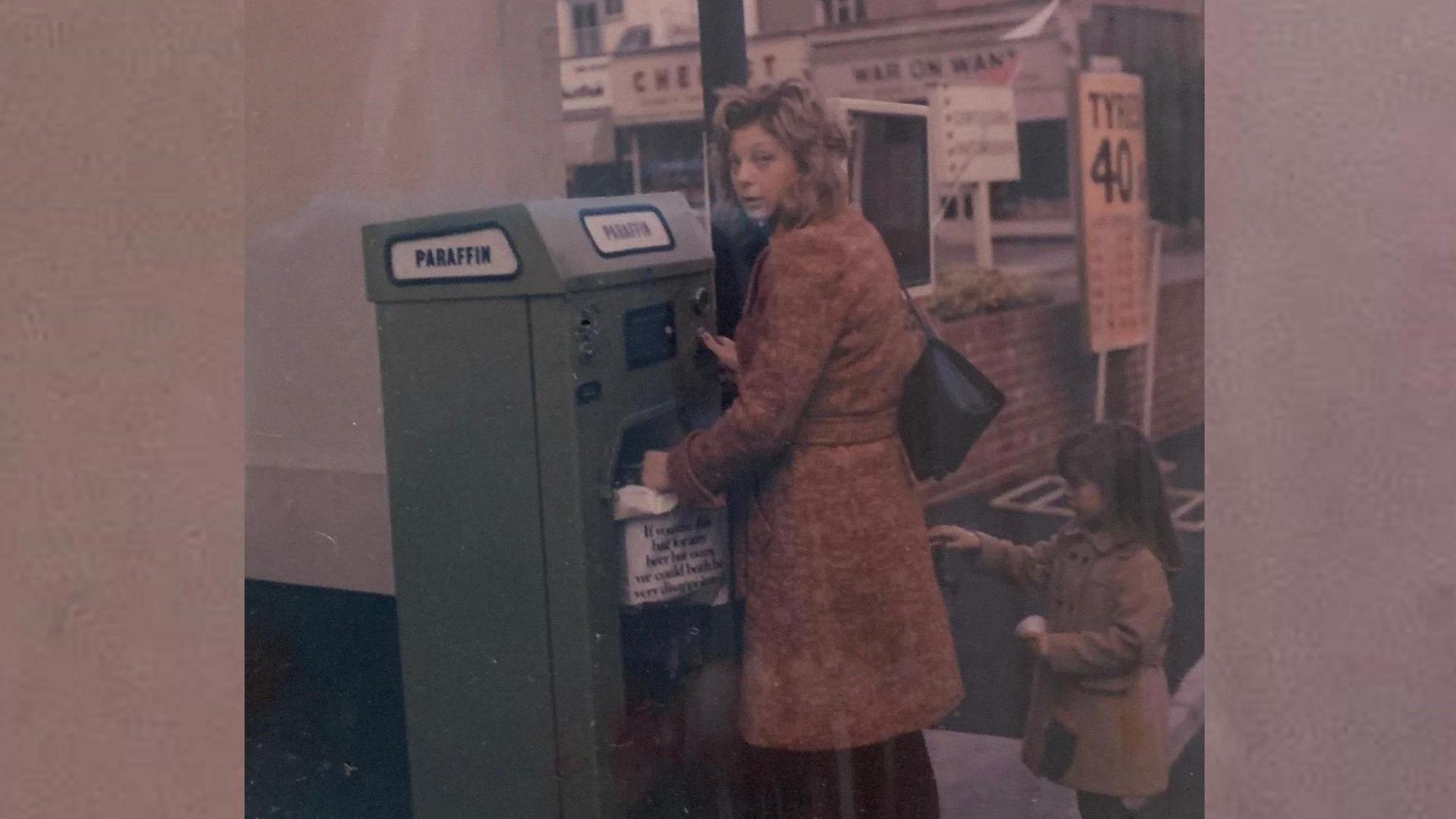 An archive photograph of a woman buying a parking ticket in the 70s. A little girl is standing behind her wearing a pink coat.