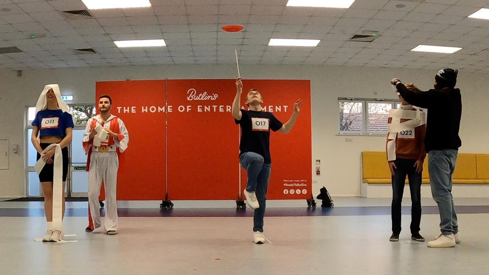 A man stands on one leg spinning a plate into the air while two pairs of people flank him while wrapping toilet roll around one another