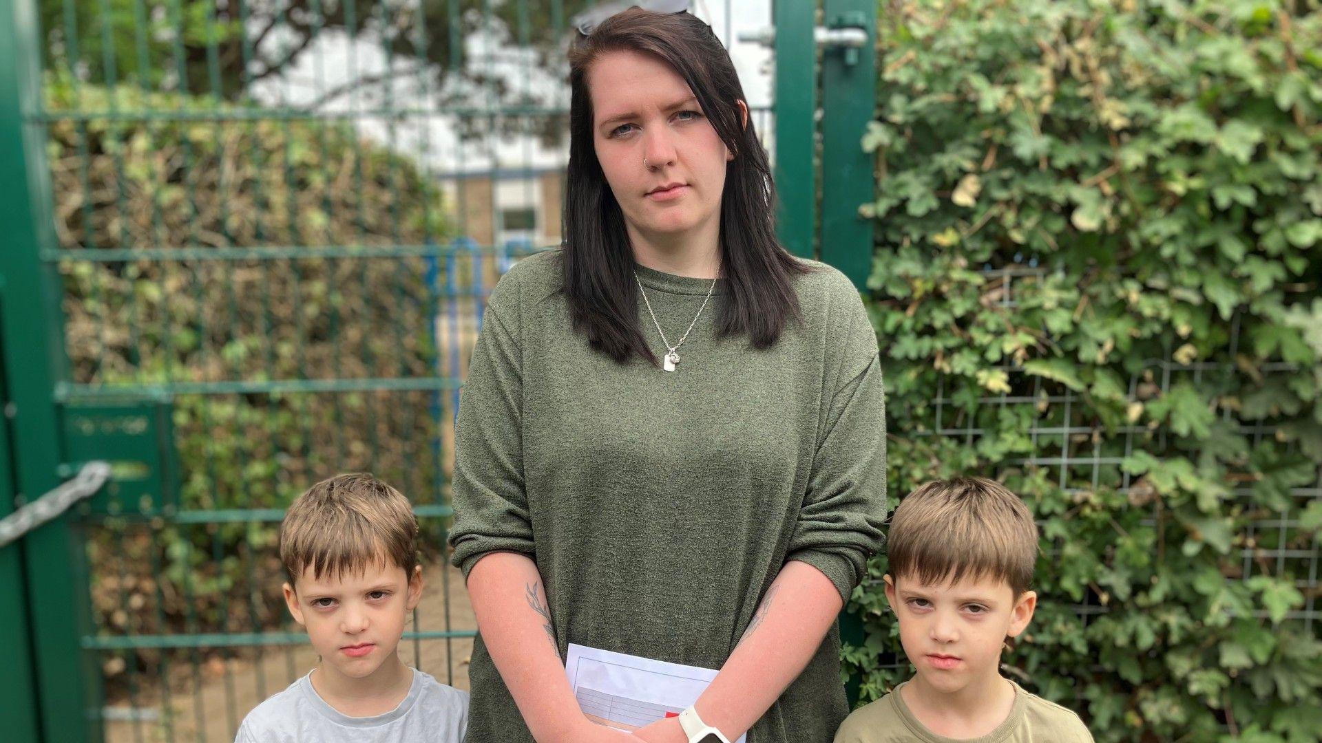 Danielle Hall and her young twin sons wear serious expressions as they stand by a fence. She has long, straight, dark brown hair and wears a green, long-sleeved T-shirt. The boys have brown hair and T-shirts.