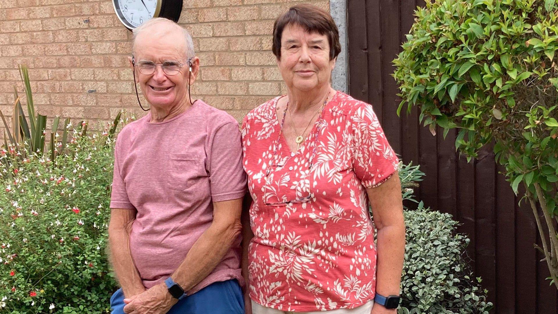 Pete wearing a pink T-shirt and blue shorts standing next to Sue wearing a pink and white floral shirt and white trousers - both standing in their back garden 
