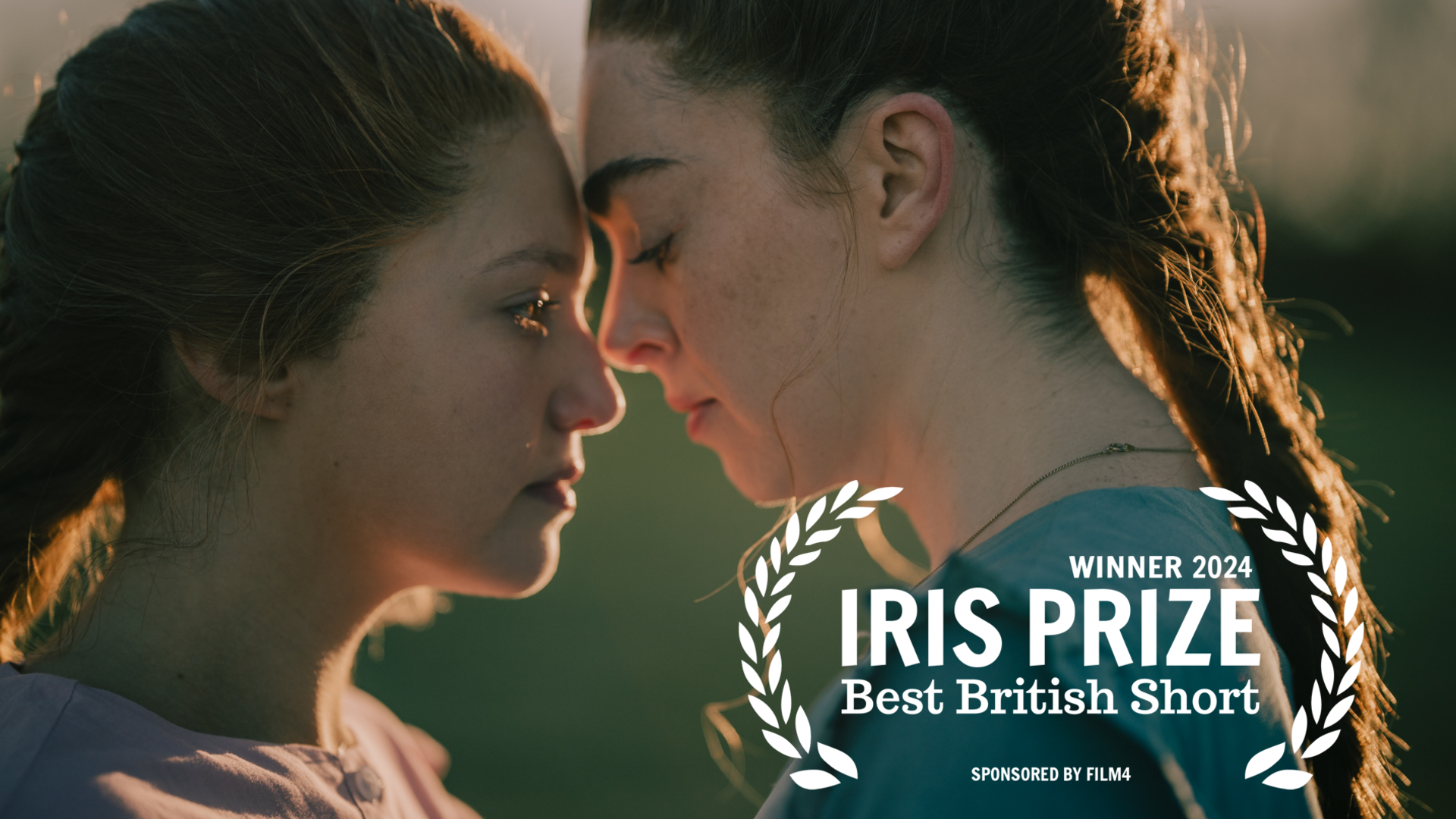 Two women in the film Sister Wives, both with serious faces, face each other and appear to touch foreheads. The picture also carries the words "Winner 2024, Iris Prize, Best British Short, Sponsored by Film4"