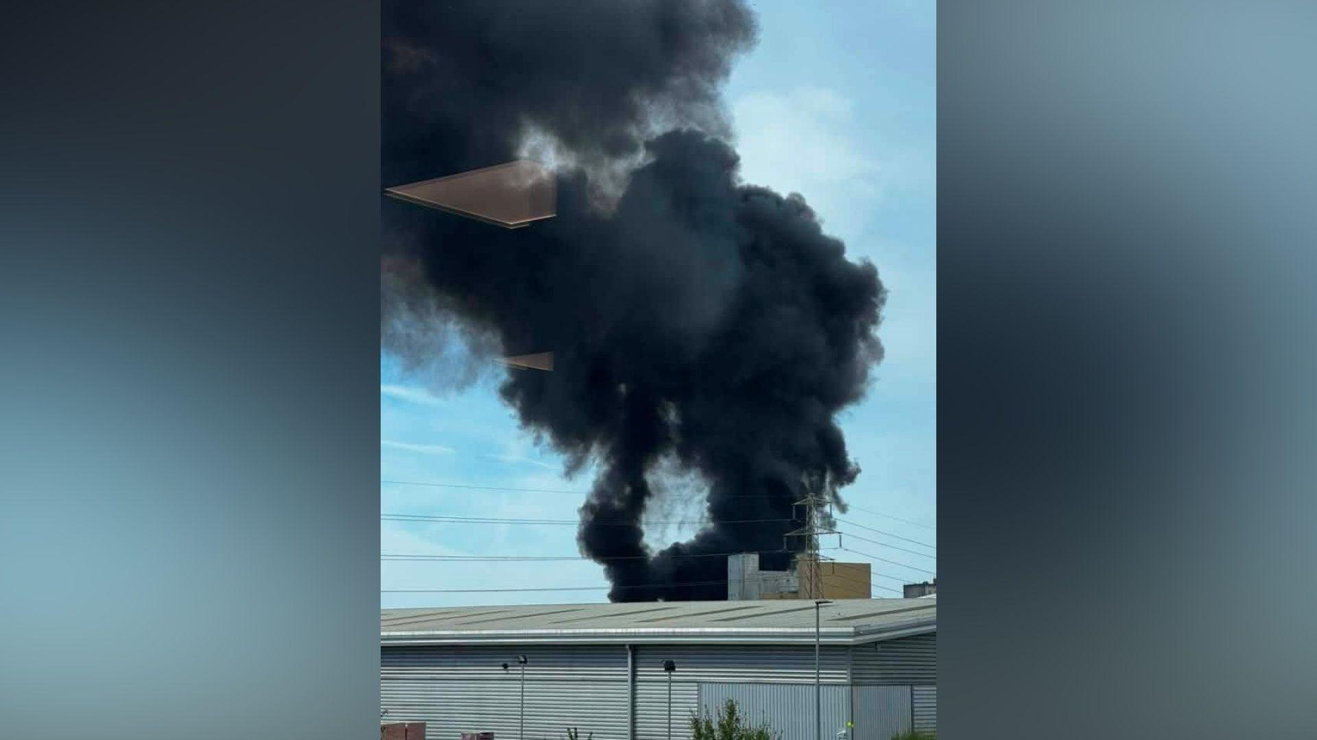 A black plume of smoke rising into the air. There is a large industrial unit in the foreground.