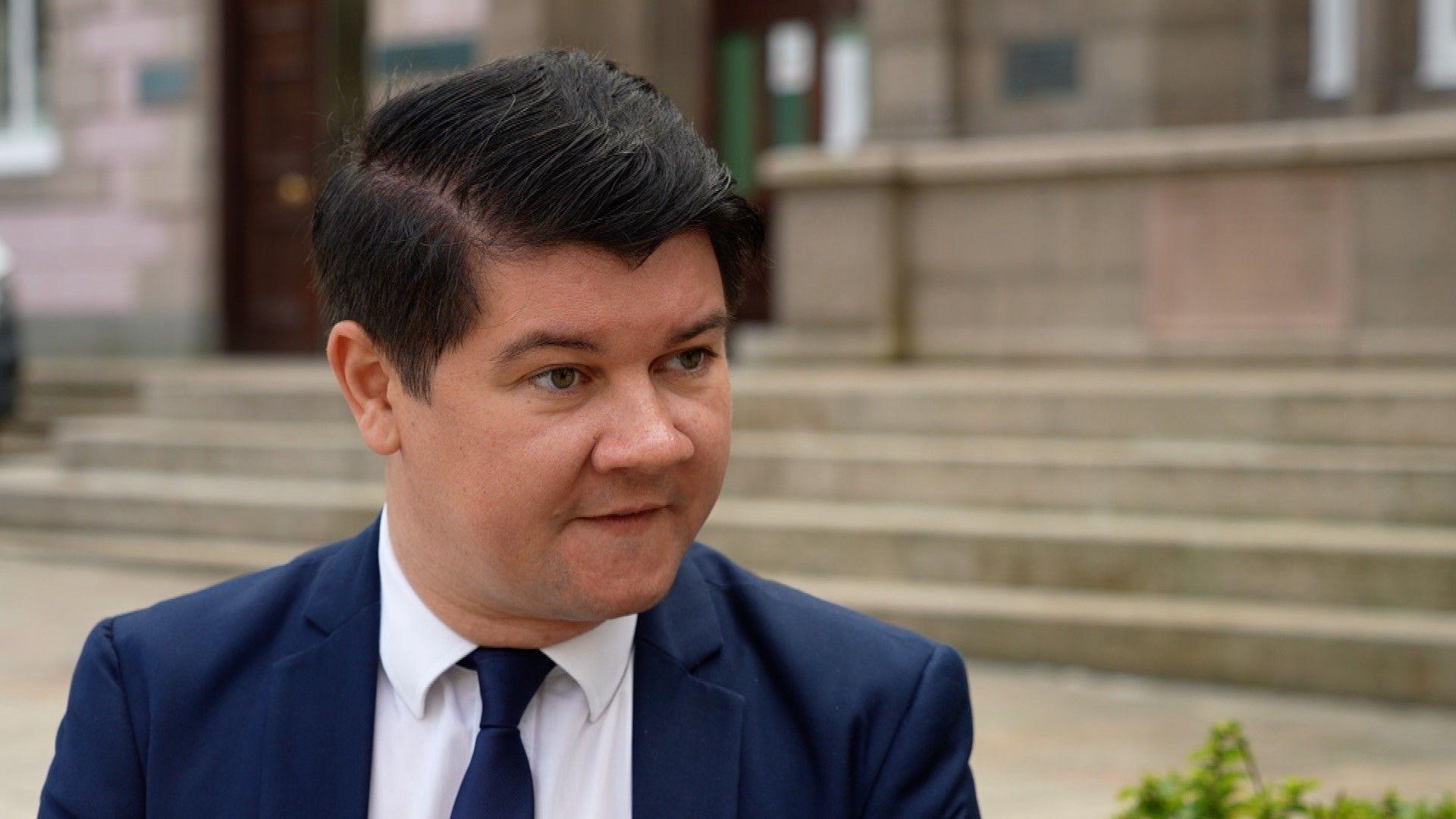 Deputy Sam Mézec in the Royal Square waring a navy suit and tie with white shirt underneath looking to the right of the frame