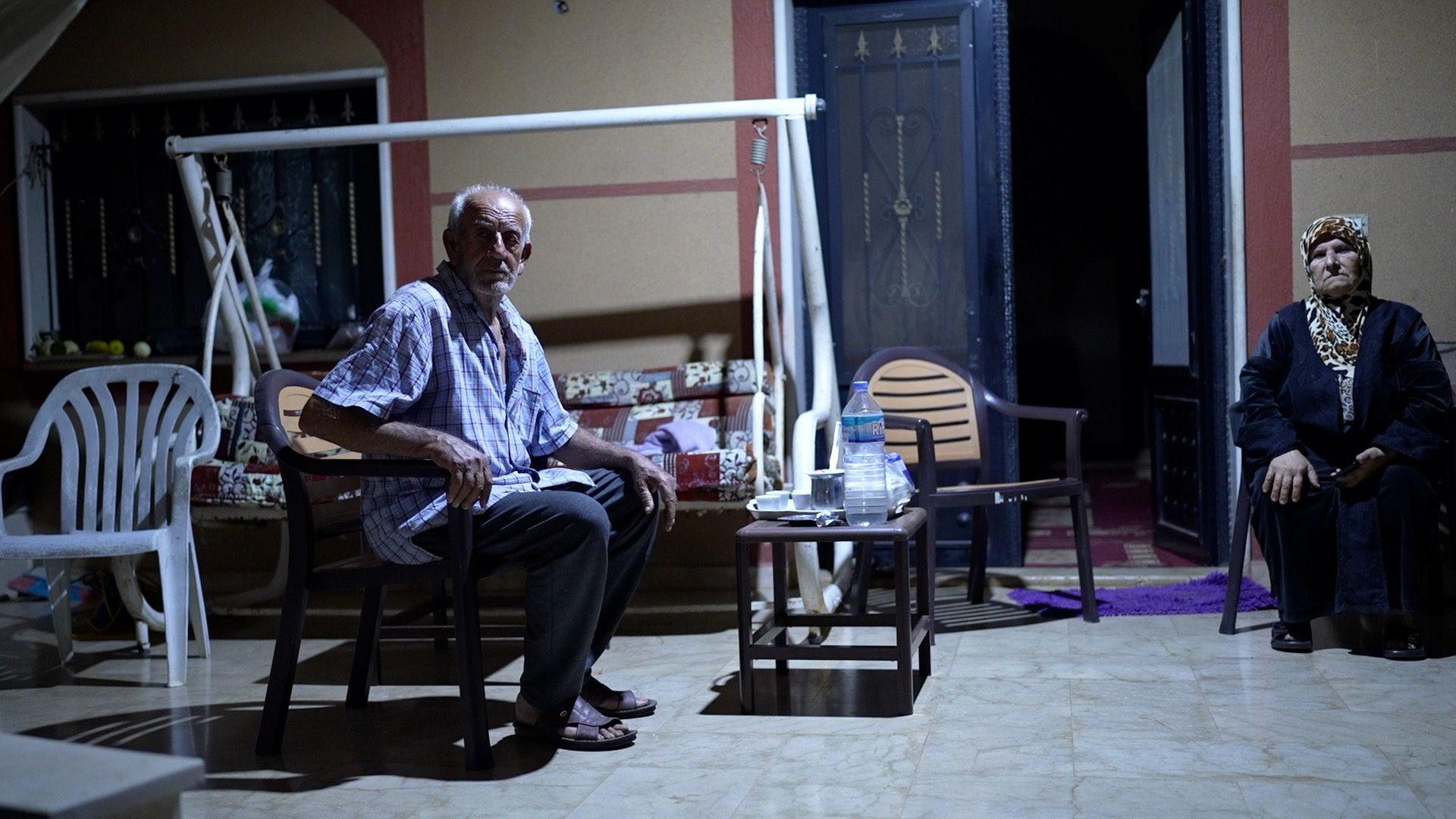 An older man and woman sit on a tiled area outside a house, after dark.