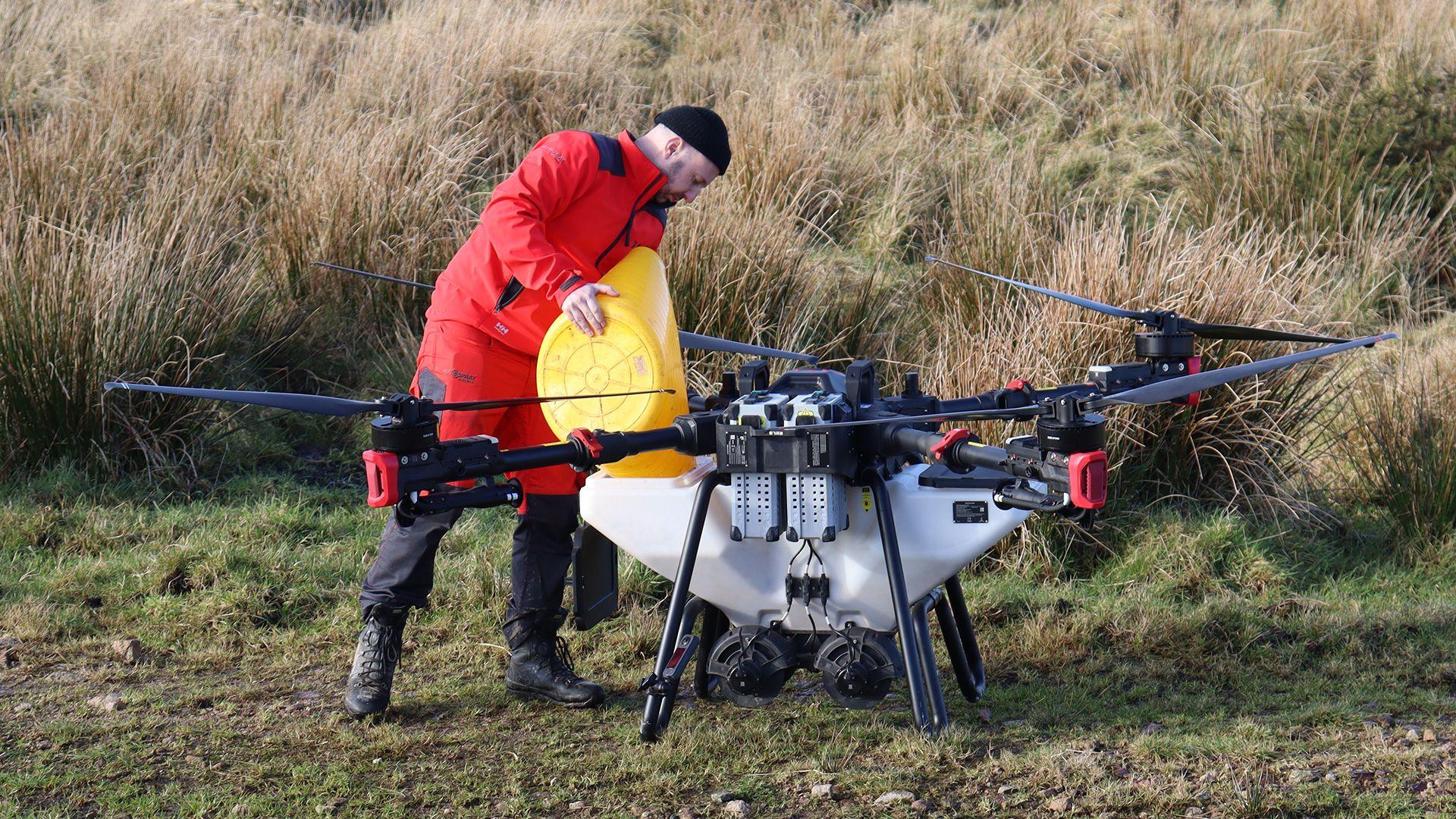 A worker in red overalls pours seeds from a yellow buckets into a large grey drone with four rotors 