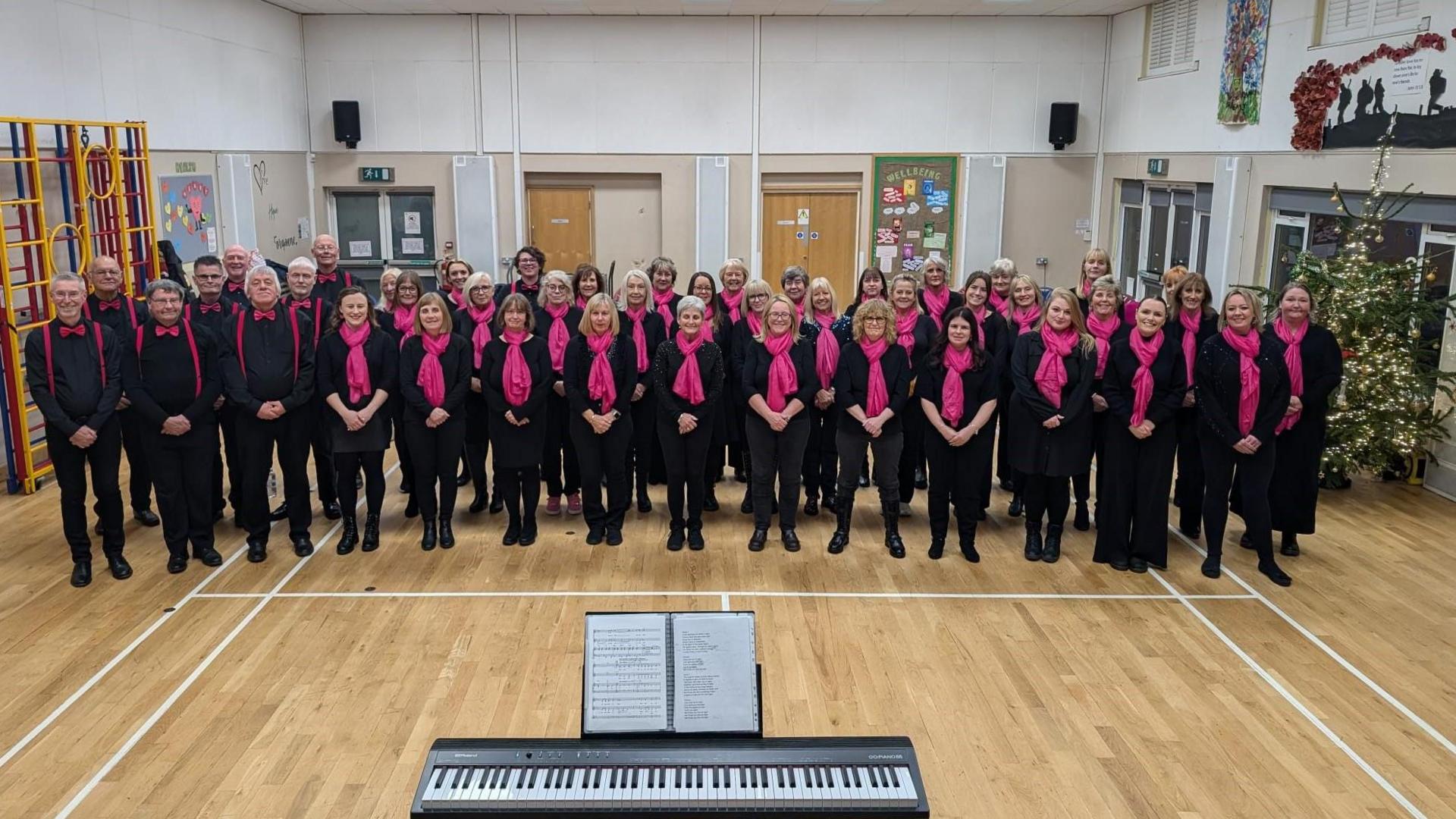 A choir of men and women dressed in black with pink scarves. There is a Christmas tree on the right hand side and a keyboard in the foreground. They are standing in a large sports hall.