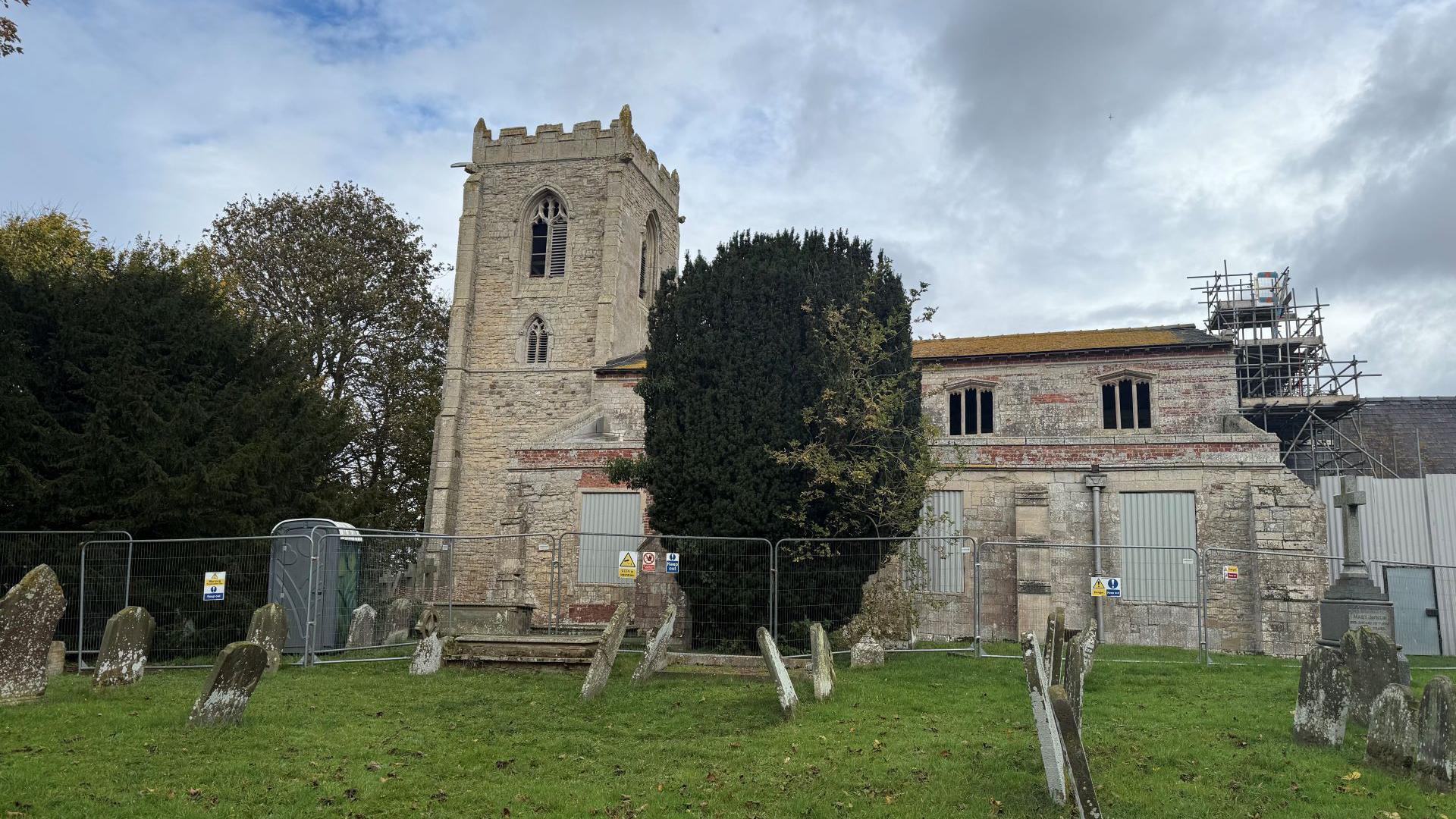 A grey stone and brick church. There are silver metal fences around the building and scaffolding to the top right. In the foreground is a burial ground. Many of the gravestones are wonky. Large green trees stand to the left, with one in the centre.