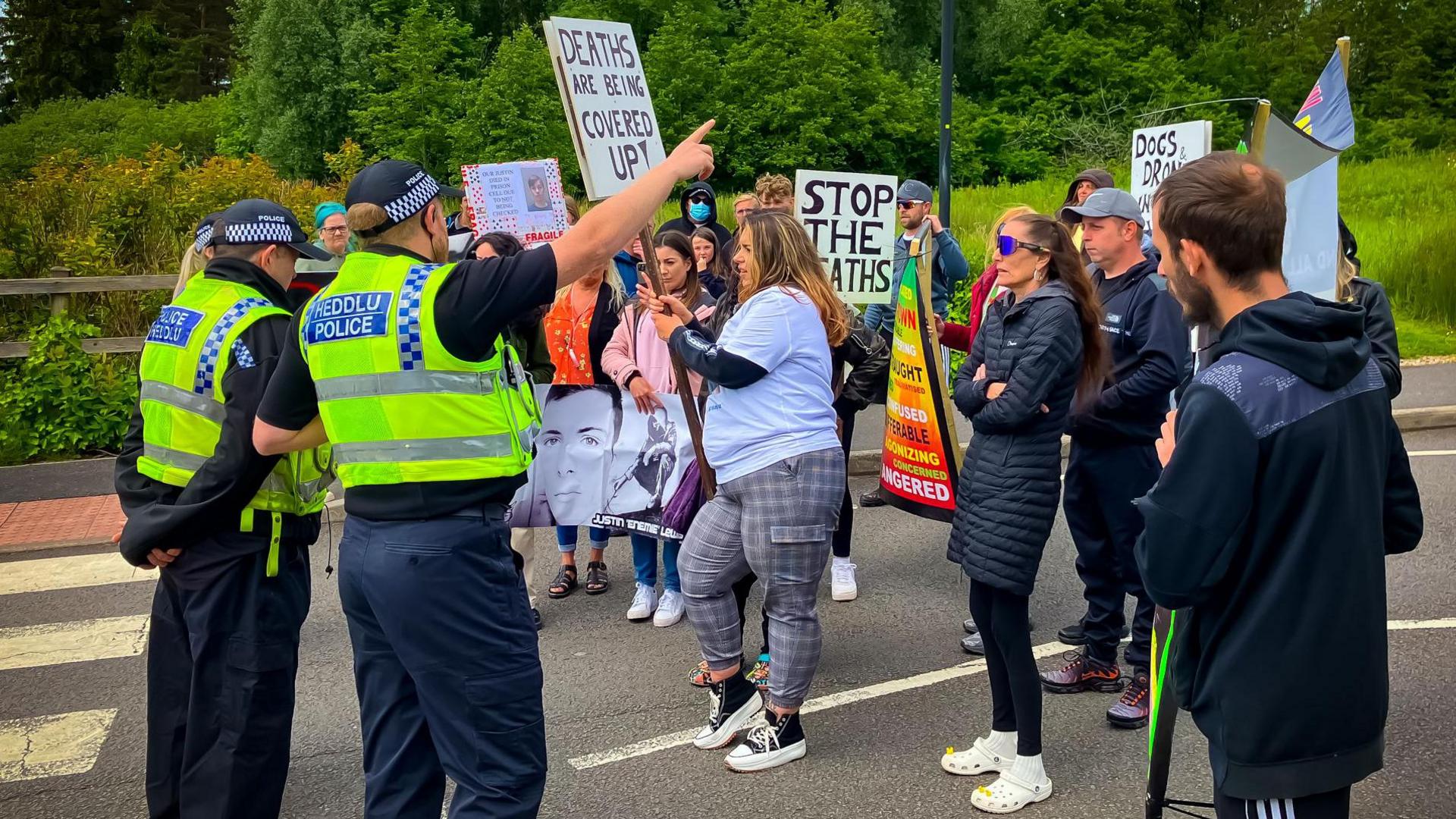 protestors holding signs saying things like "stop the deaths" stand on a road in front of three police officers