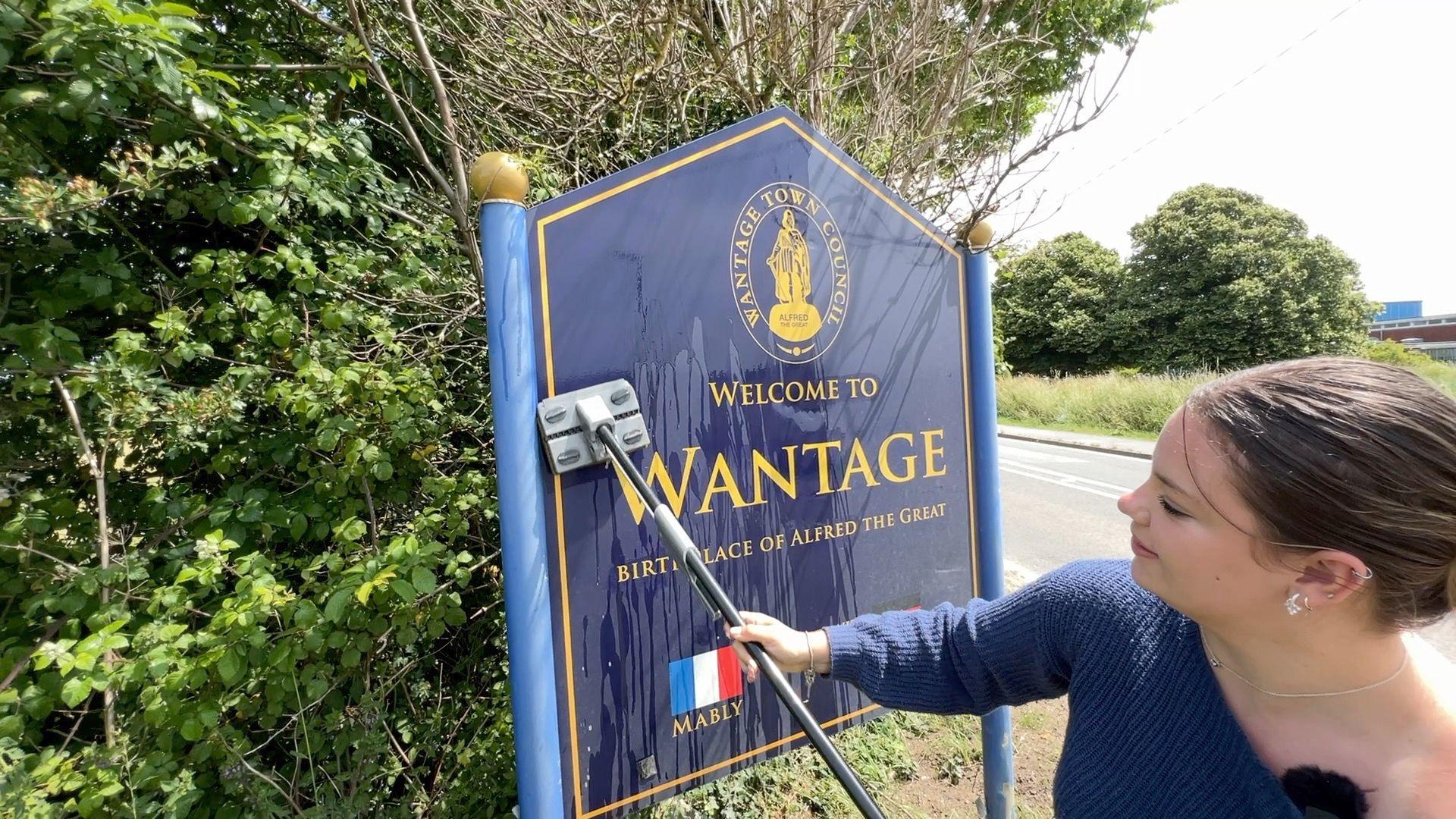 Ellie, who has brown hair tied-up, is using a mop to clean a large blue road sign welcoming motorists in to Wantage.