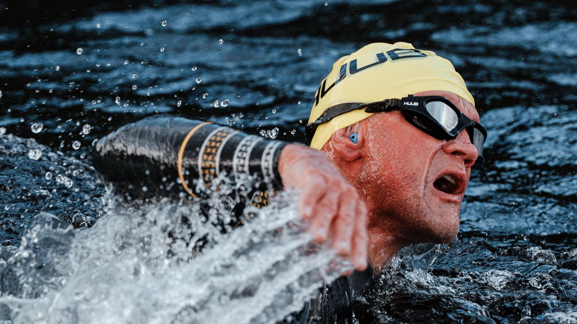 A close-up of Iain Sinclair as he swims through choppy, dark blue water. He is taking in a breath as he raises an arm from the water. He is wearing a black wet suit, yellow swim cap and swimming goggles.